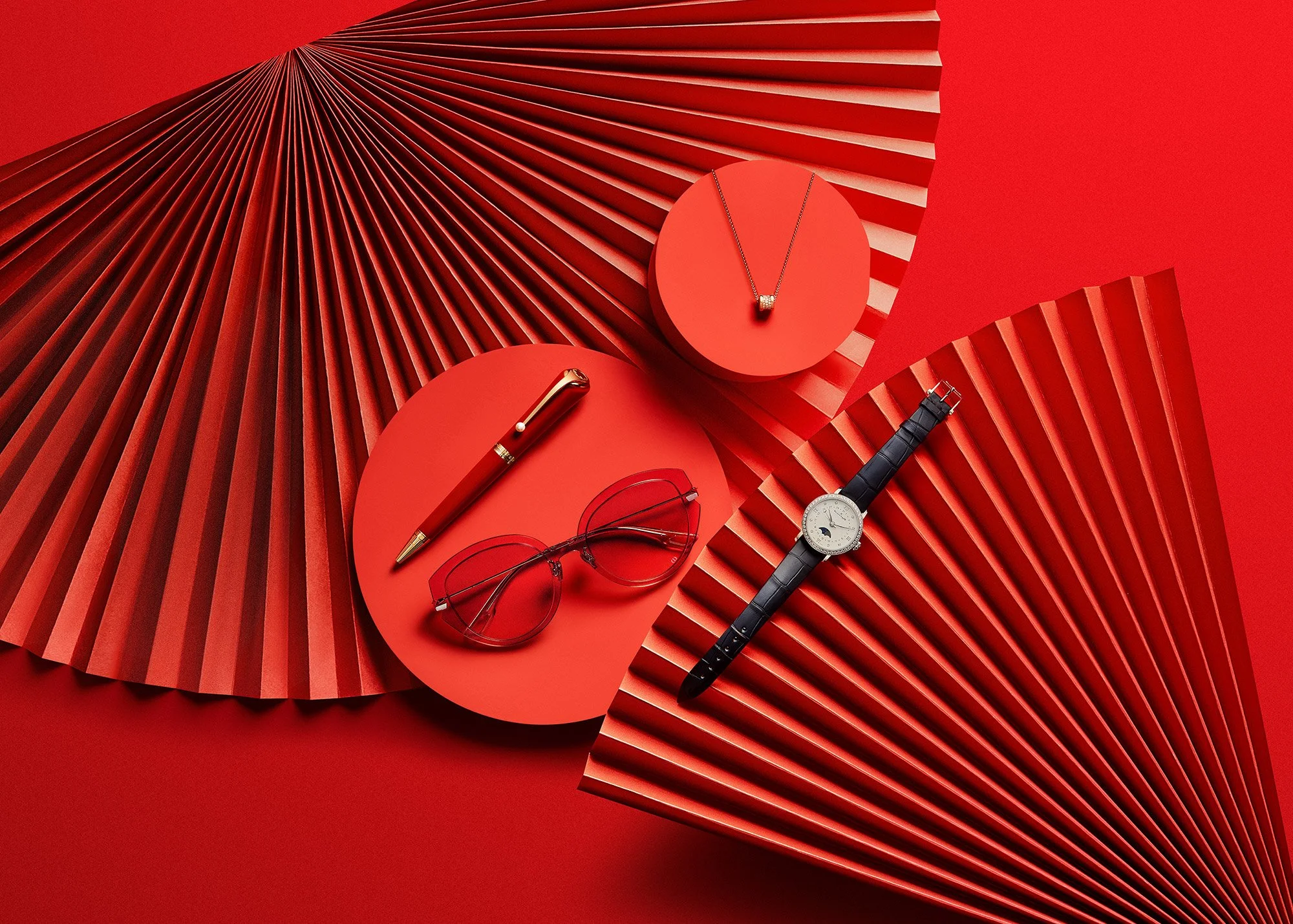 Red background with red pleated paper and platforms displaying accessories: a gold wristwatch, a necklace with a pendant, a pair of red glasses, a red pen, and a black leather strap watch.