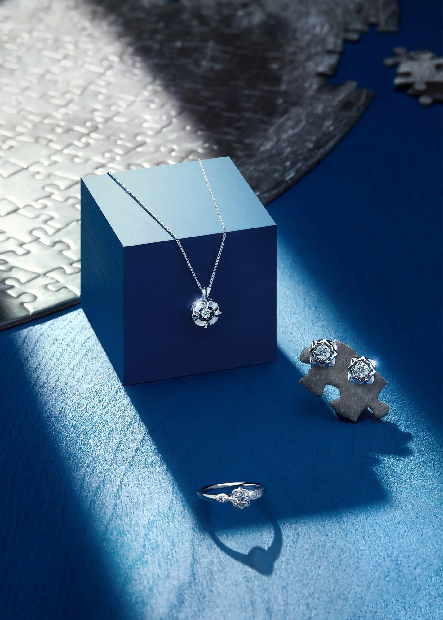 Jewelry set including a necklace with a diamond pendant, a pair of diamond earrings, and a diamond ring, all displayed on a blue surface with puzzle pieces and decorative elements in the background.