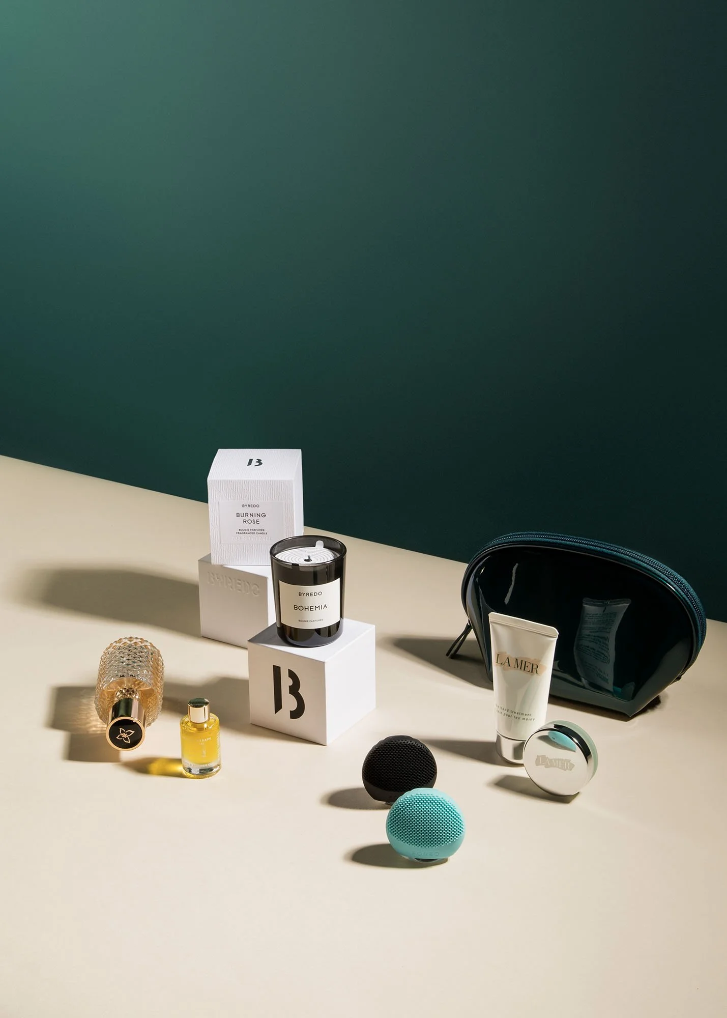 Collection of beauty and skincare products, including a candle, small perfume bottles, facial creams, and a blue makeup bag, arranged on a flat surface with a green and beige background.