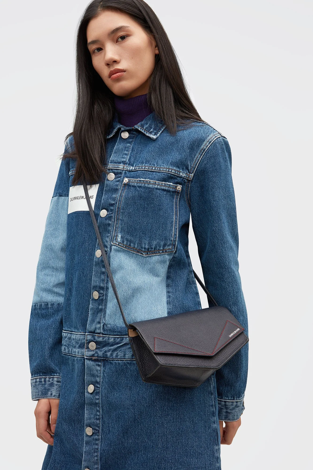 A woman with straight black hair wearing a denim jacket with a Calvin Klein Jeans label, a purple turtleneck, and carrying a black leather crossbody bag with red stitching.