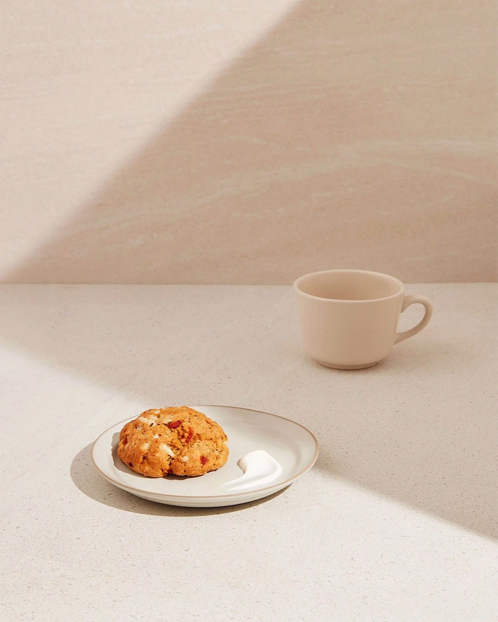 A plate with a cookie and a dollop of cream, a white mug, and a textured, light-colored surface with a shadowed background.