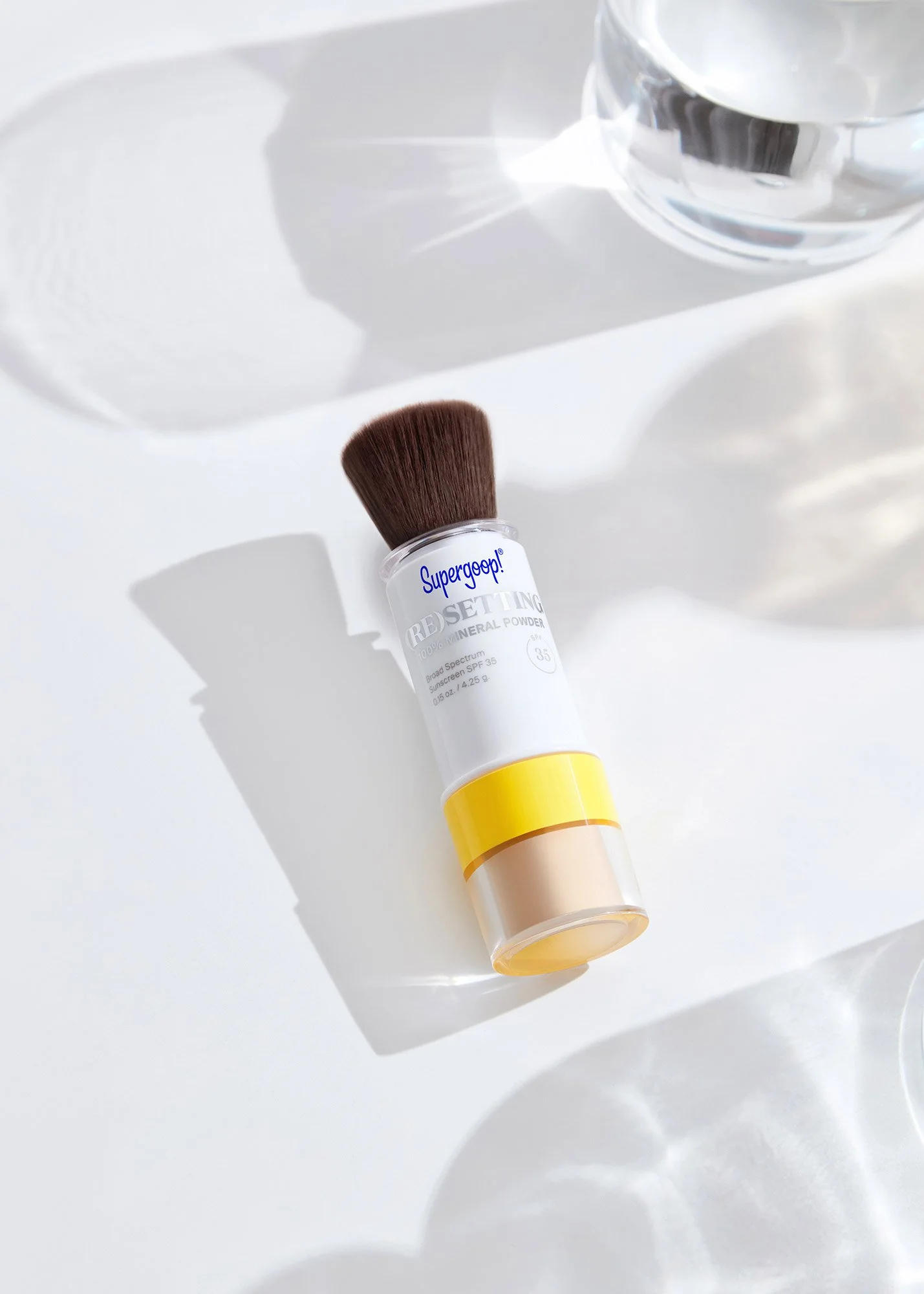 A makeup brush with a yellow and clear handle and a brown bristle head rests on a white surface. There is a glass of water nearby, casting reflections.