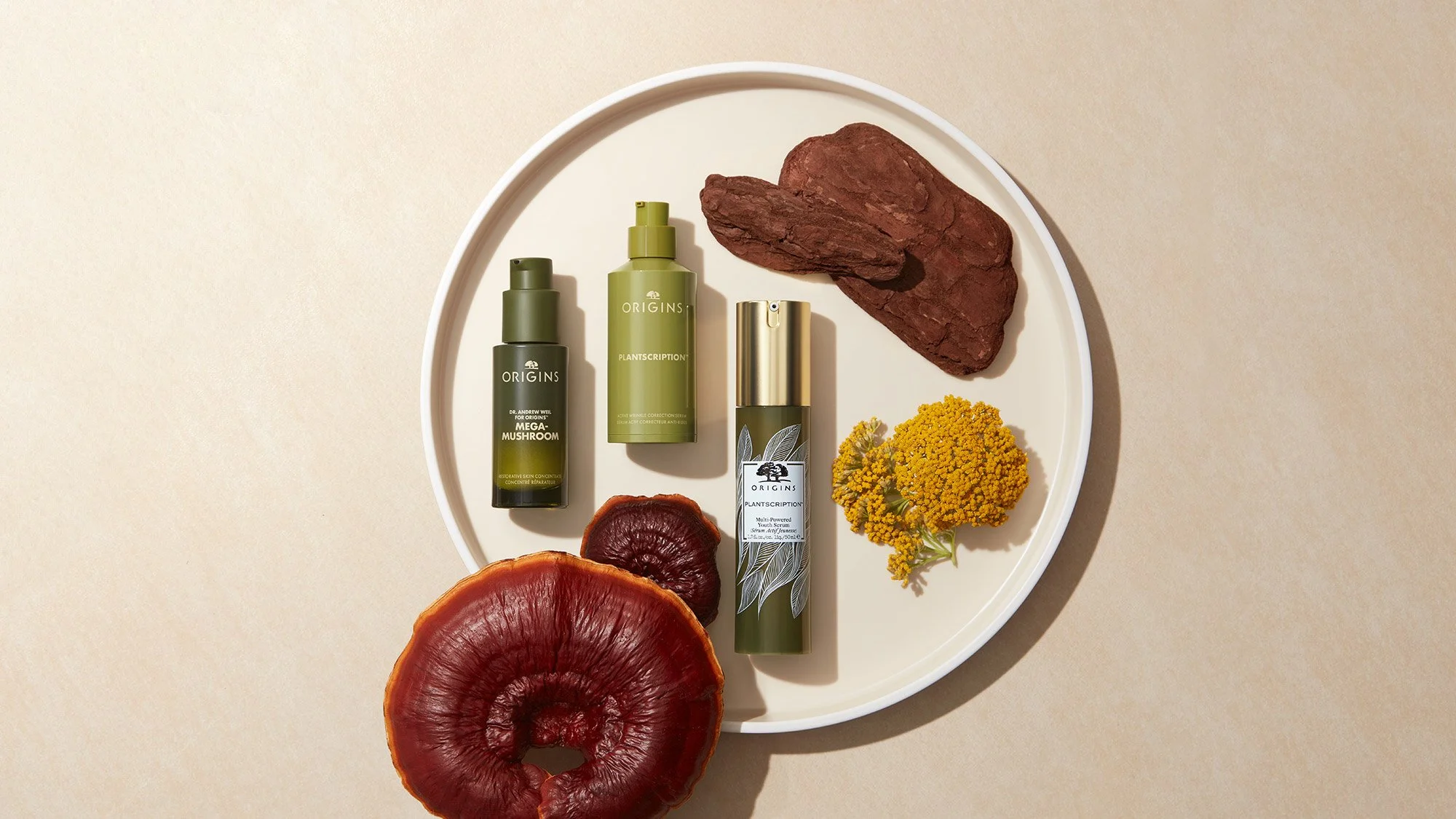 A white circular tray with skincare products, dried red mushrooms, yellow flowers, and brown bark placed on a beige background.