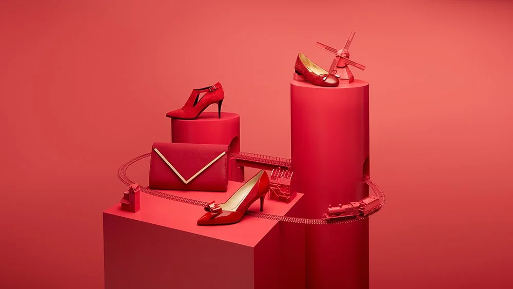 A red-themed display of women's fashion accessories, including high heels, a clutch purse, and miniature models of a house and train, with a red train track sculpture looping around the display
