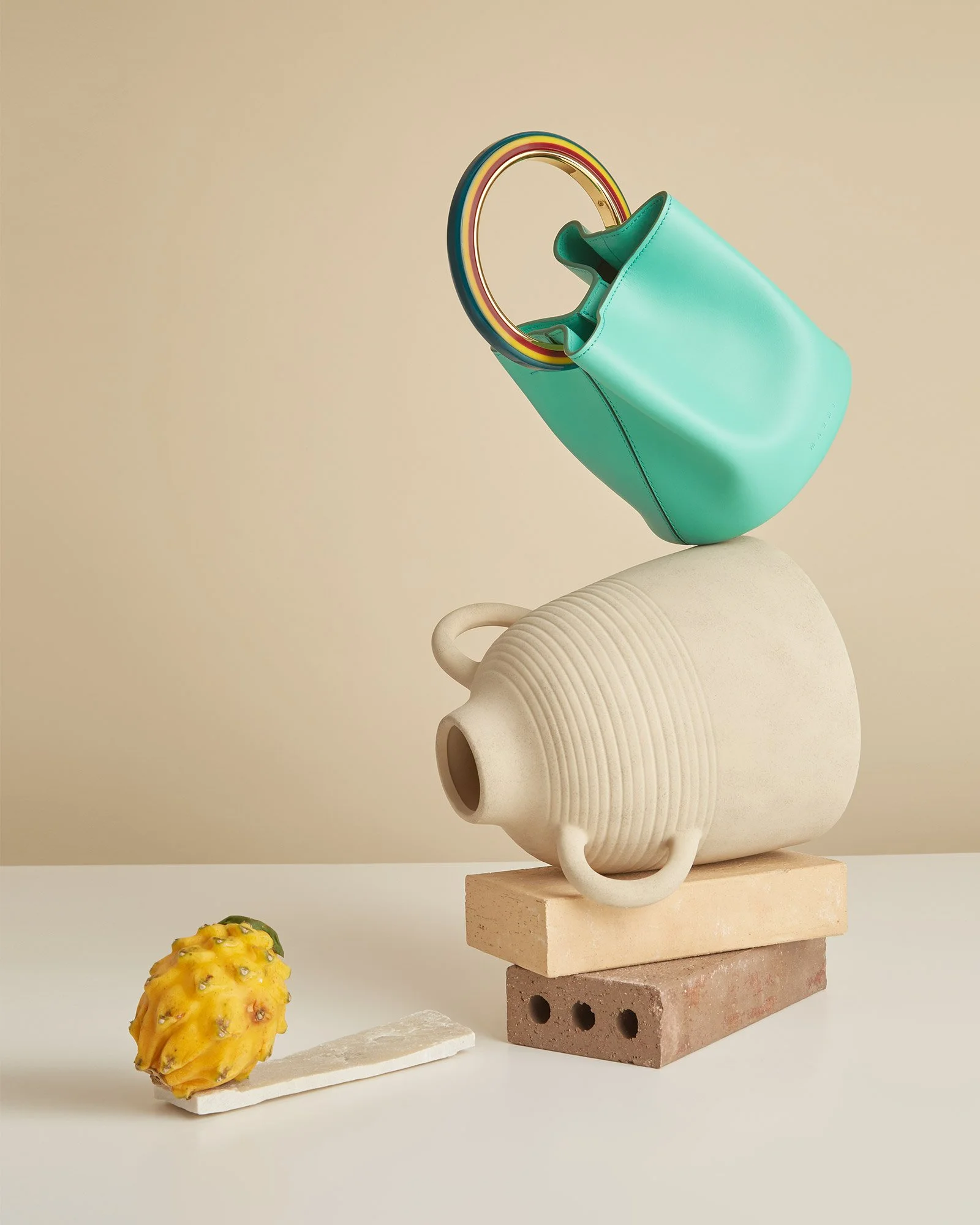 A turquoise handbag with rainbow-colored handles balanced on top of a white ceramic vase with circular ridges, which is placed on top of layered bricks. A small yellow fruit or decorative object is on the white surface in front.