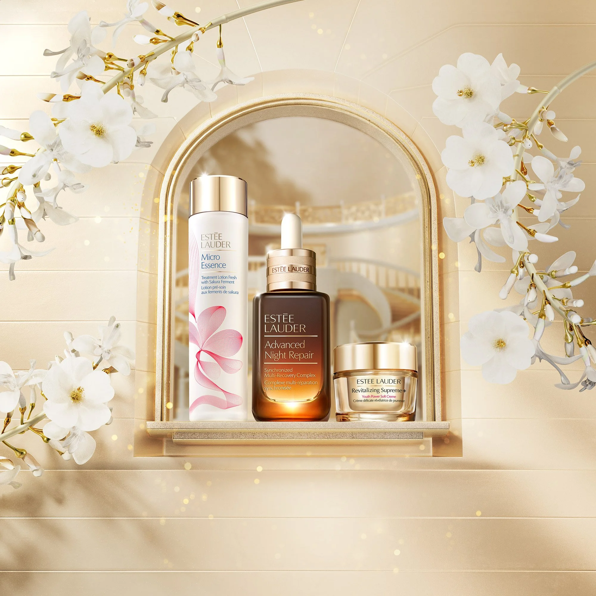 Three Estee Lauder skincare products on a shelf in front of a mirror, with white and gold flowers around the window opening.