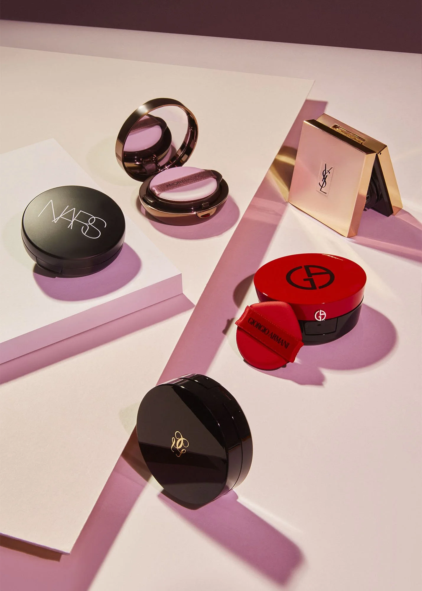 Several luxury makeup compacts and cases, featuring brands like NARS, Yves Saint Laurent, Giorgio Armani, and Giorgio Armani in a pink-themed display with soft lighting.