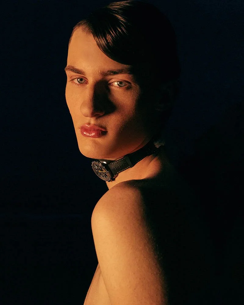 A portrait of a young person with short dark hair, wearing a black choker with a round pendant, looking over one shoulder in dim, warm lighting.