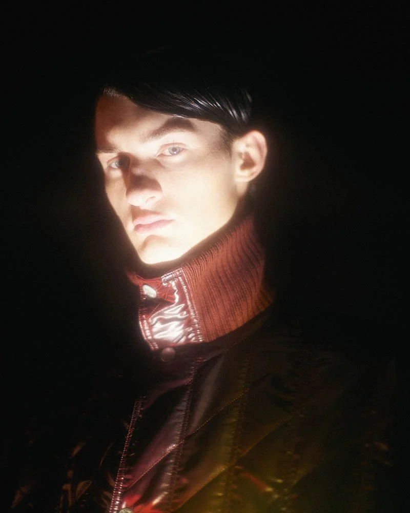 A person with short dark hair, wearing a red jacket with a high collar, is illuminated against a dark background, mostly showing their face and upper torso.