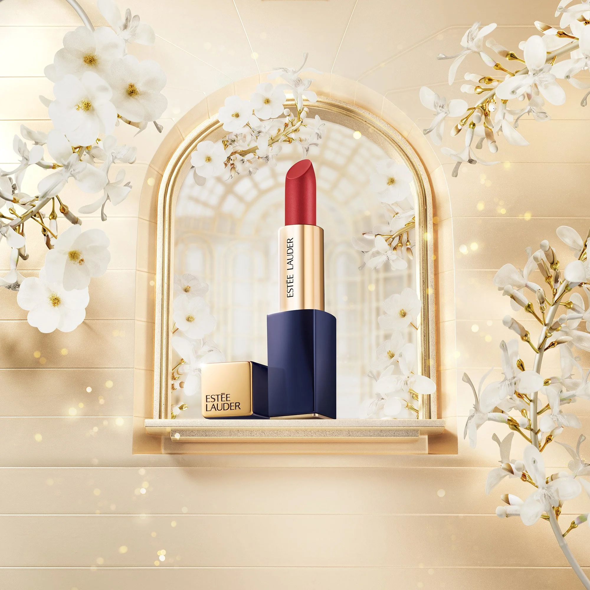 A lipstick from Estée Lauder with a gold and navy blue case, positioned on a marble shelf in front of a mirror with a gold frame. Surrounding the lipstick are white flowers and gold accents, creating an elegant, luxurious setting.