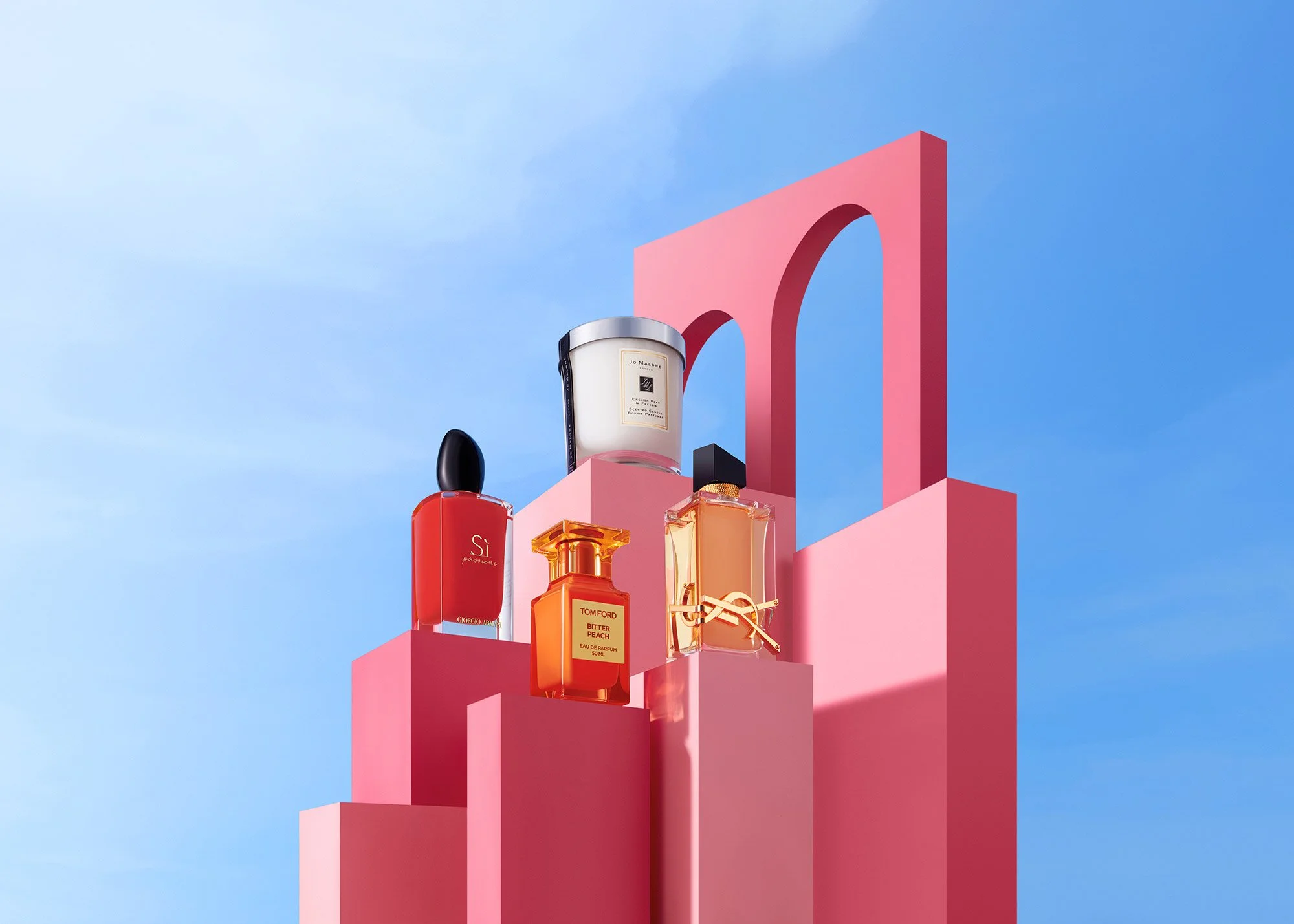 A display of five perfume bottles on pink tiered platforms against a blue sky with scattered clouds.