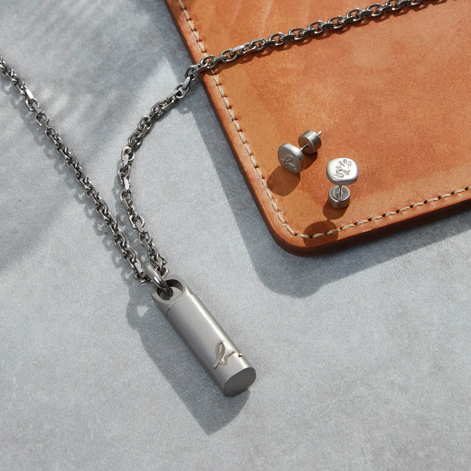 A silver necklace with a rectangular pendant engraved with a cursive letter 'L'. The necklace is resting on a light gray surface next to a brown leather piece. There are also two small silver stud earrings with engraved designs placed on the leather 