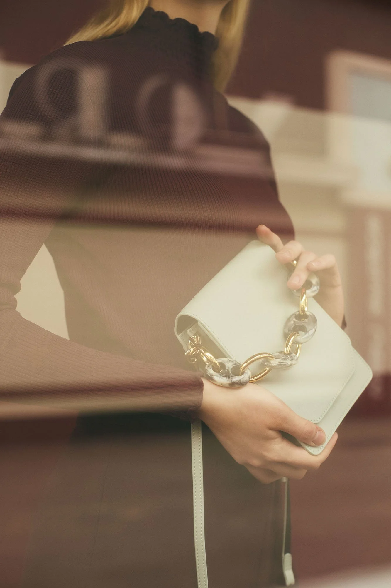 A woman in a brown sweater holding a white clutch purse with large chain links, standing in an elevator with reflective metallic walls.