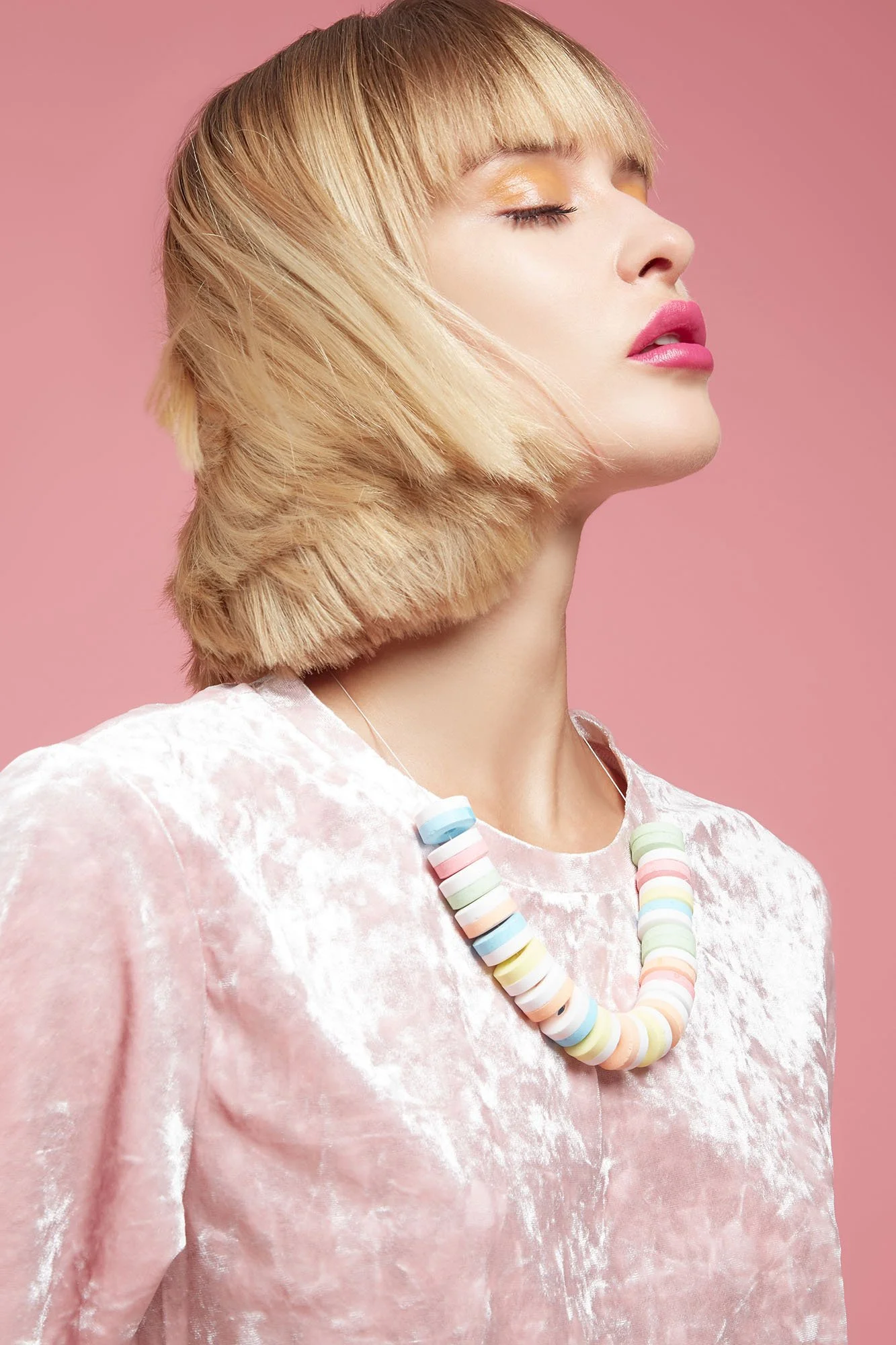 A woman with blonde hair and bangs, wearing pink lipstick, closed eyes, and a pastel beaded necklace, against a pink background.