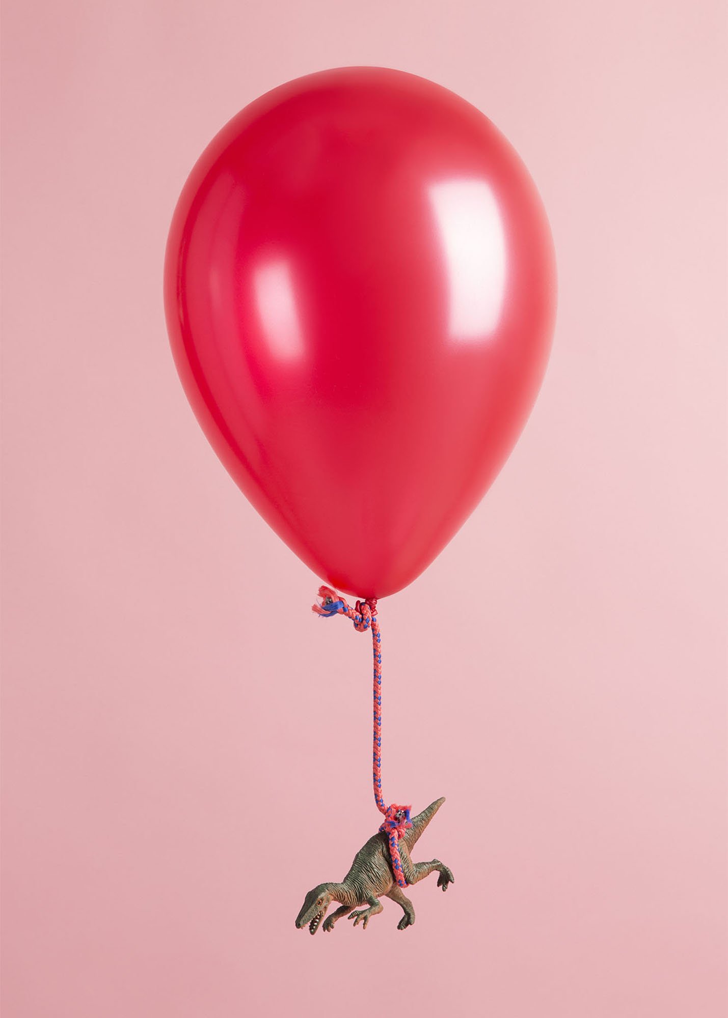 A red balloon with a string tied to a small toy Tyrannosaurus rex, set against a pink background.