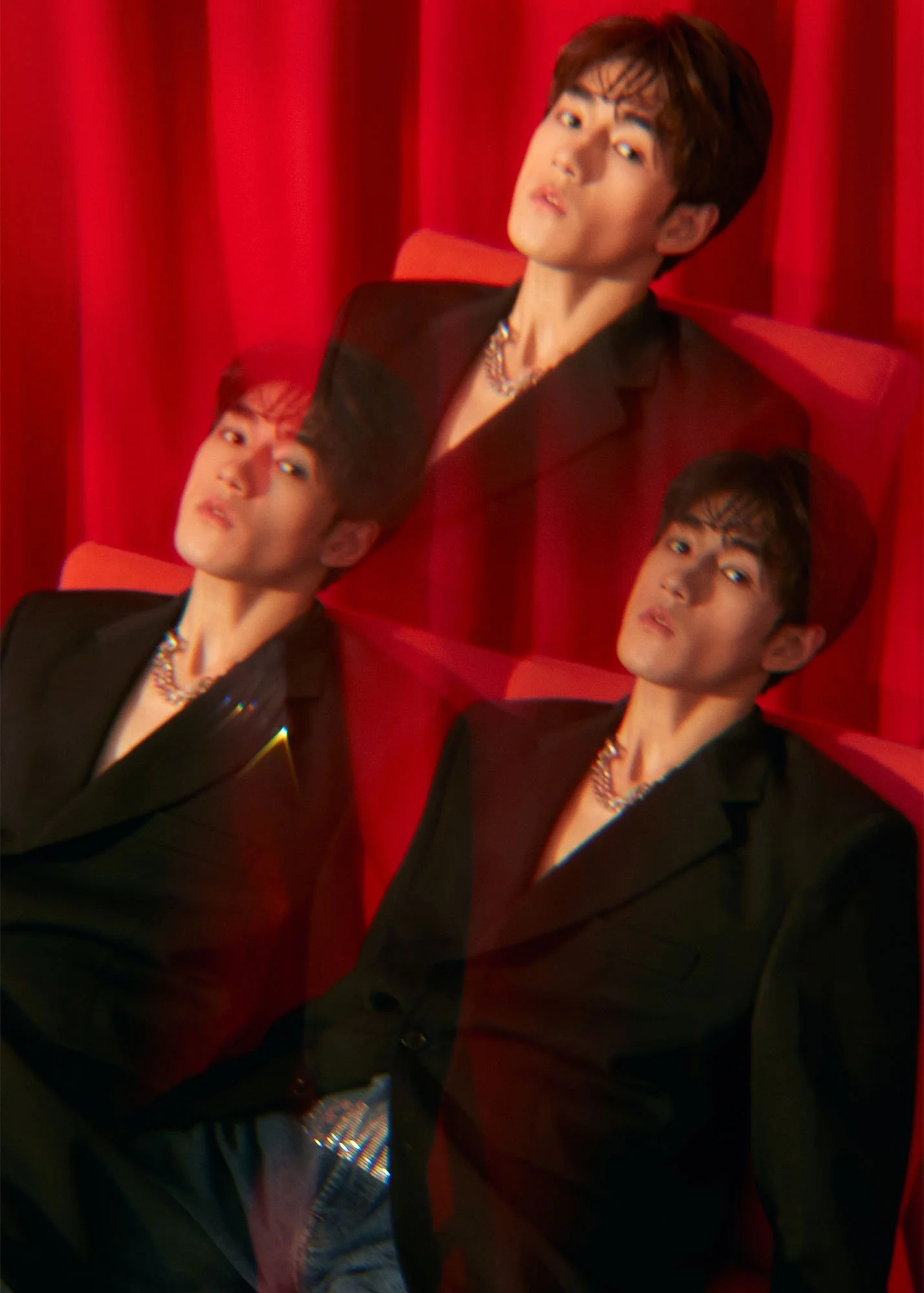 A person with short dark hair and makeup, wearing a black blazer and necklaces, sitting on red chairs against a red curtain, with a mirrored reflection of their face.