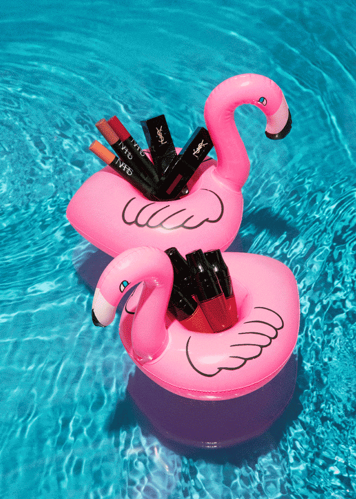 Pink inflatable flamingo drink holder floating in a pool filled with water, holding various lip glosses and makeup tubes.