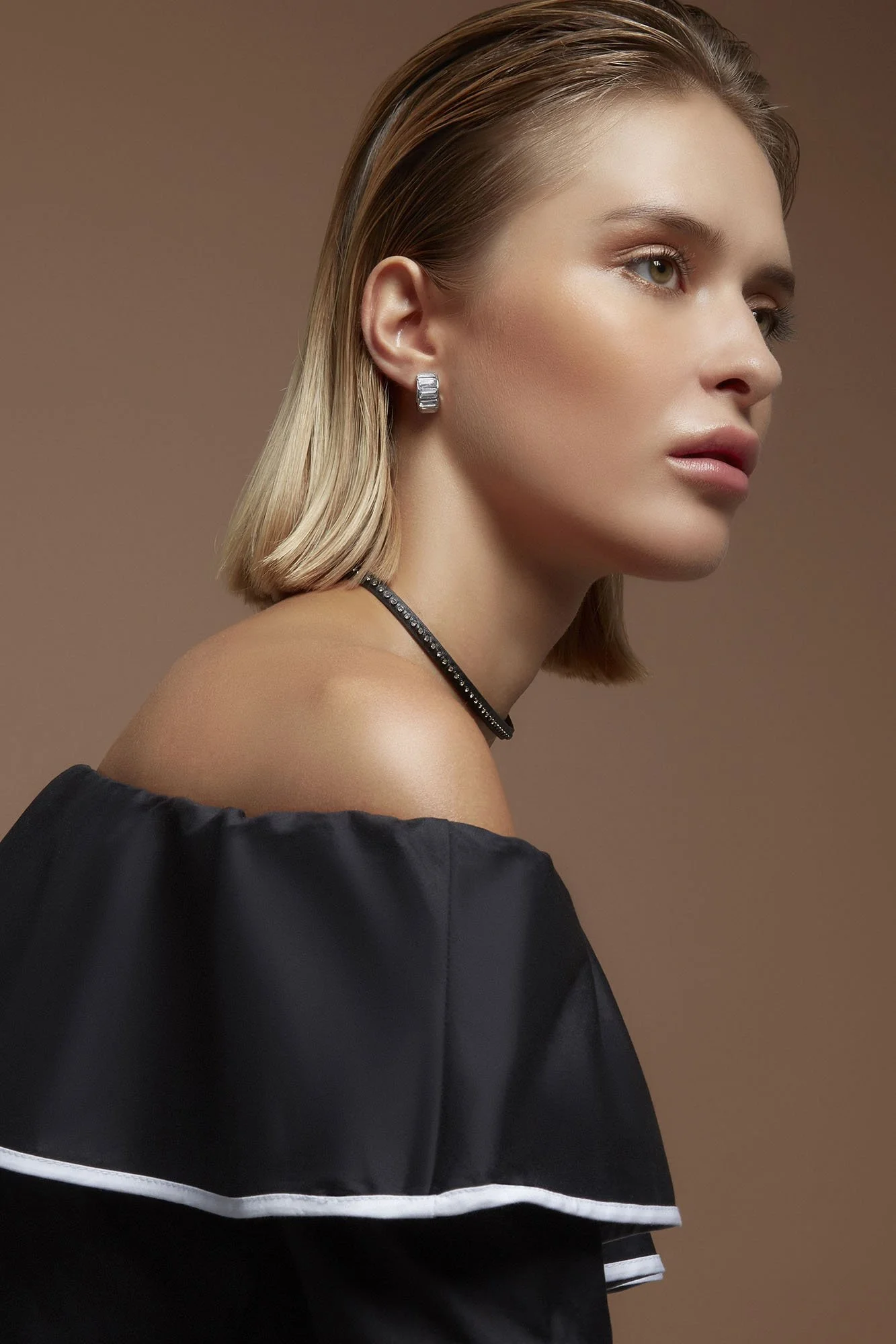 A young woman with blonde hair styled in a shoulder-length bob, wearing silver hoop earrings, a black off-the-shoulder top with a white stripe detail, and a black choker necklace with small embellishments, posed against a plain brown background.