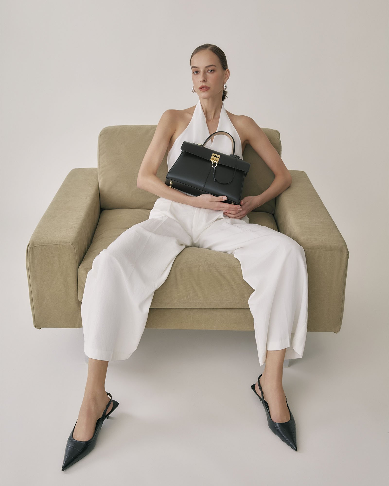 A woman with light skin, dressed in white, sitting on a beige sofa, holding a black handbag on her lap, wearing black high-heeled shoes, with her legs apart and arms resting on her thighs, against a plain light background.