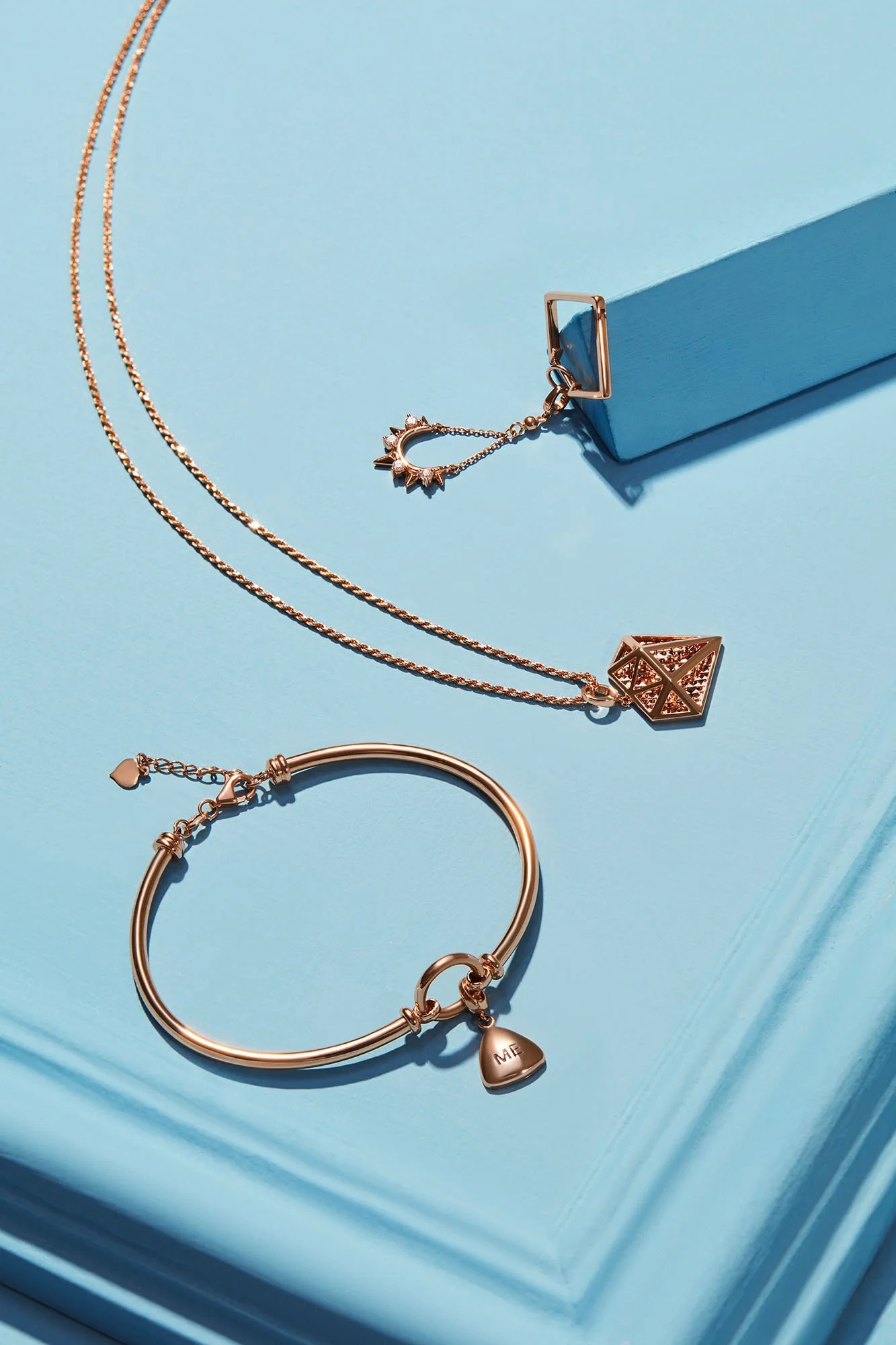 Gold jewelry including a necklace with a geometric pendant, a bracelet with a 'ME' charm, and a chain with a star and sun motif, displayed on a blue surface with a blue box.