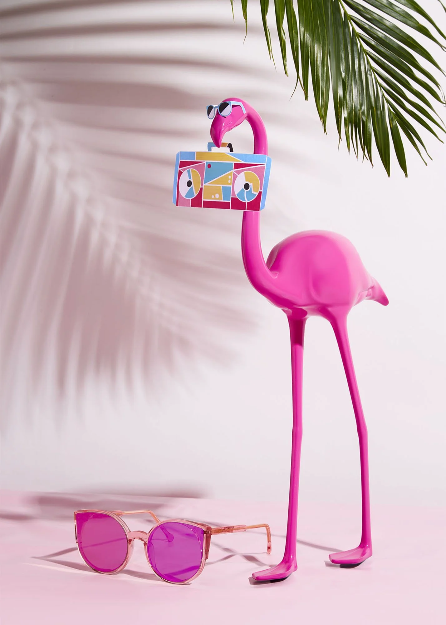 Pink plastic flamingo with sunglasses, holding a colorful boombox, pink sunglasses on the ground, and a palm leaf in the background.