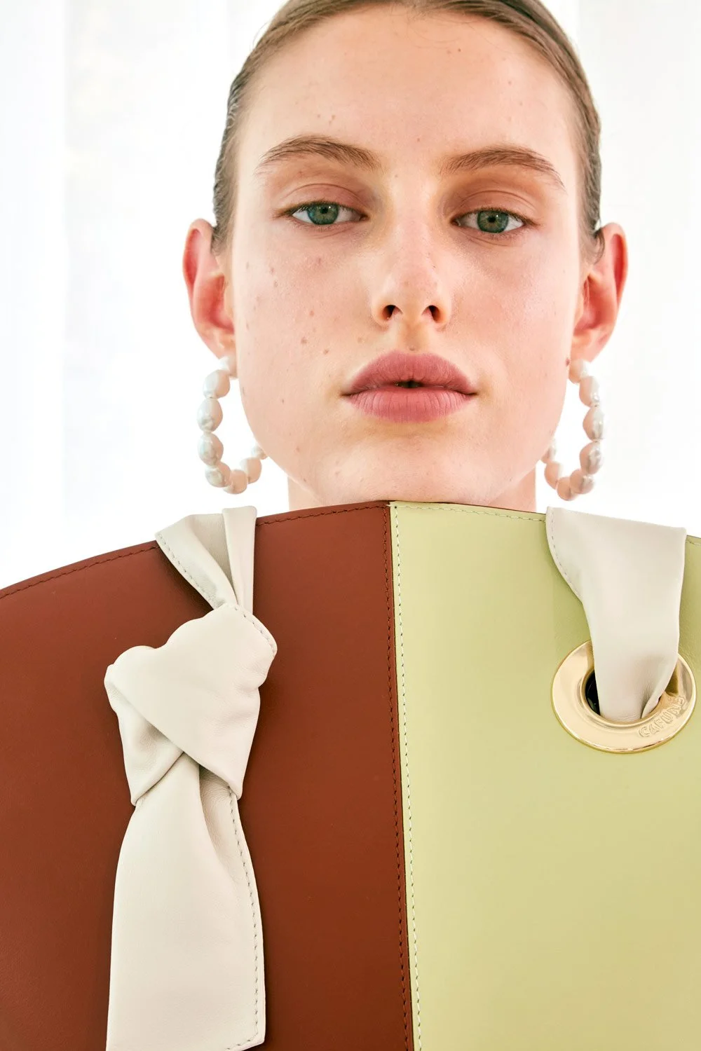 A woman with light skin, green eyes, and blonde hair, wearing large pearl earrings, holding a large, multicolored bag with a cream bow, photographed against a white background.