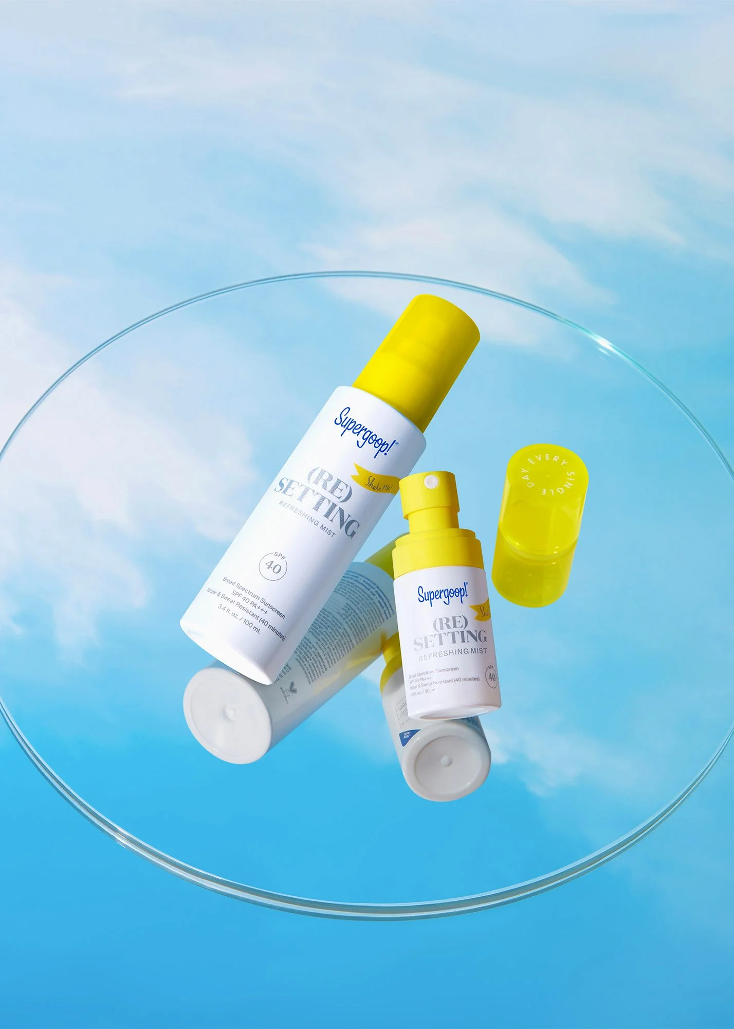 A clear glass tabletop with three bottles of Supergoop! skincare products, including a refreshing mist with SPF 40, set against a sky background with clouds. The bottles are white with yellow caps and labels.