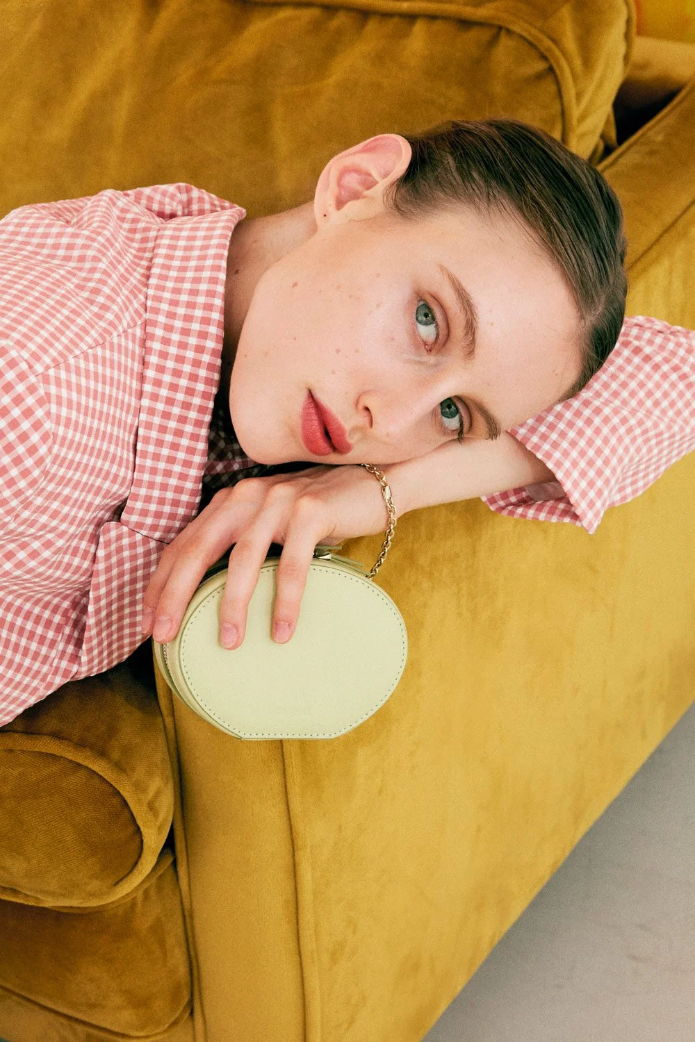 A woman with blue eyes and short dark hair lies on a mustard-colored couch, resting her head on her arm and holding a yellow handbag with a chain strap. She's wearing a pink and white checkered shirt and has a relaxed expression.