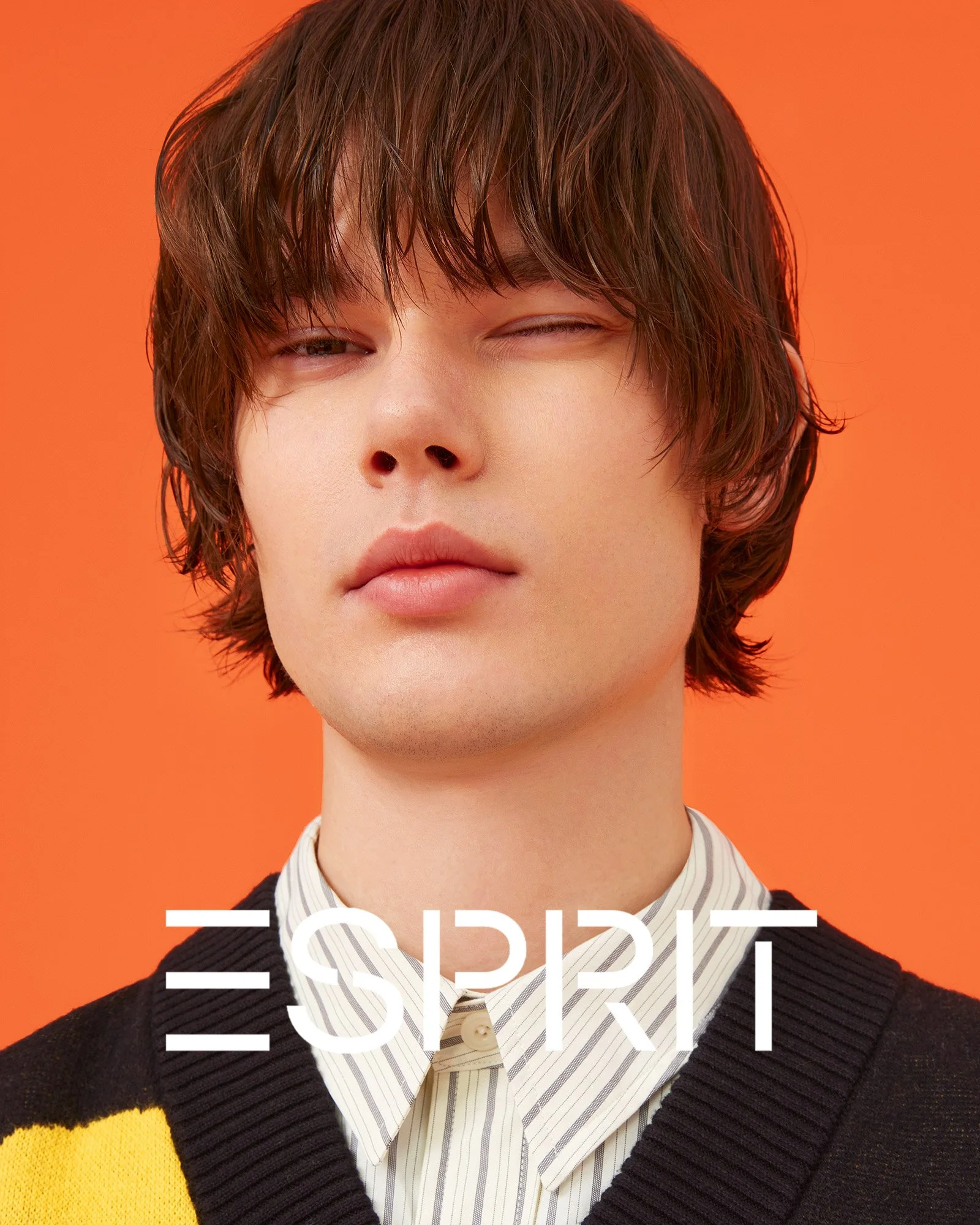 A young man with brown hair in a layered hairstyle, wearing a striped shirt and a dark sweater with yellow accents, posing against an orange background, with the brand name 'ESPRIT' overlaid.
