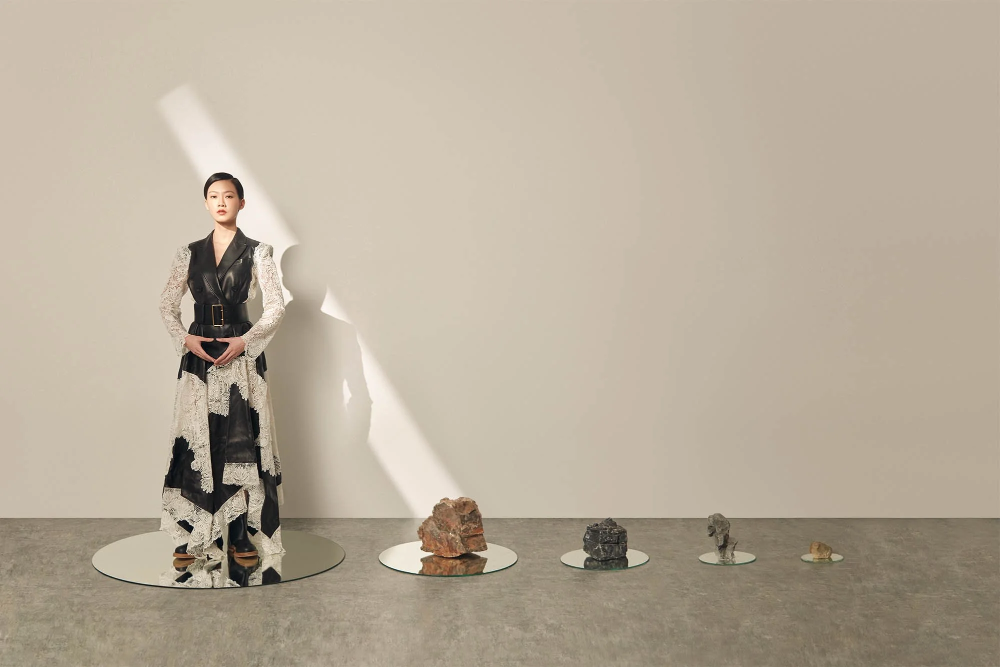 A woman standing on a circular mirror with smaller mirrors and rocks arranged in a row on the floor, in front of a beige wall. She wears a black and white lace dress with leather details.