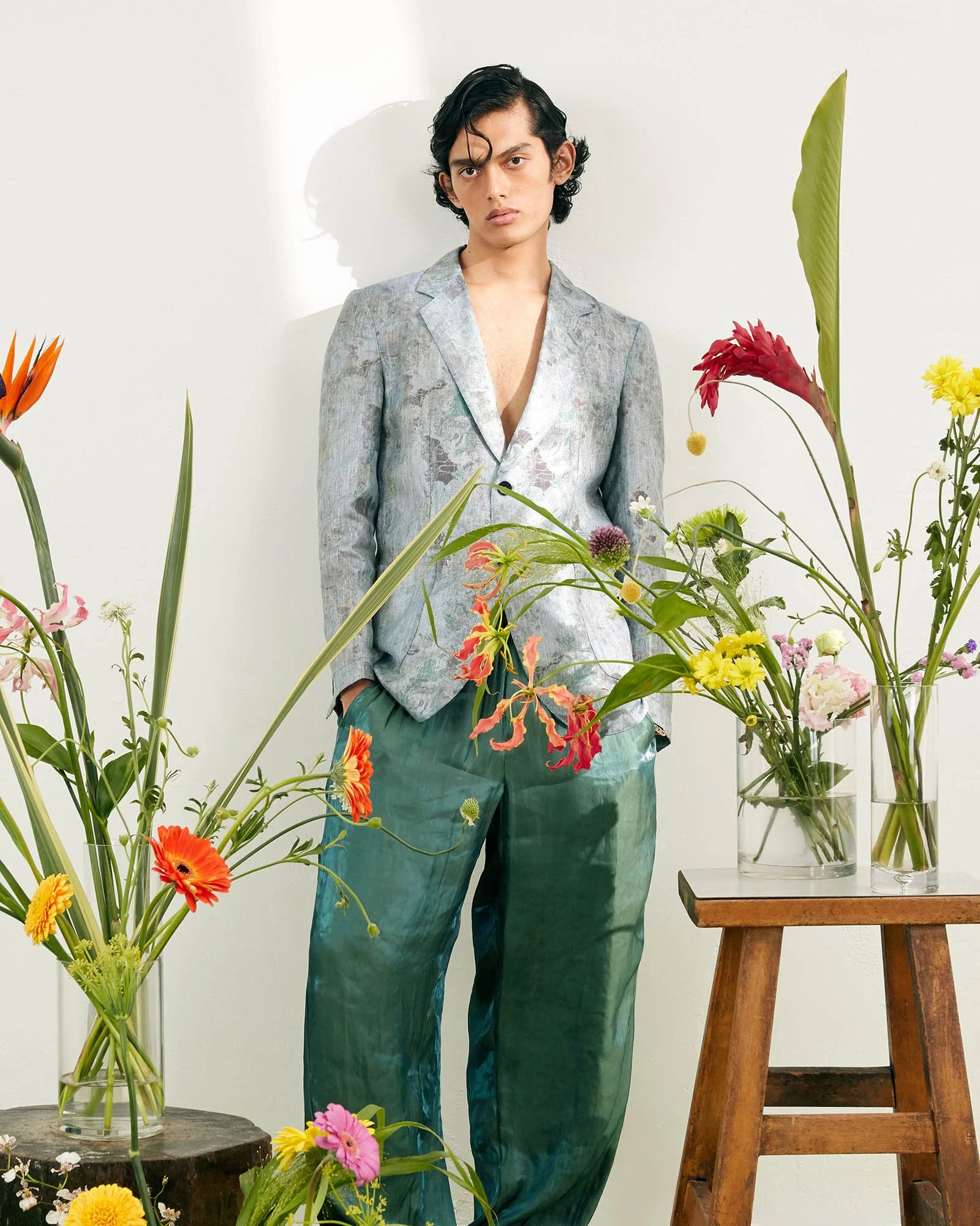 A young man with dark, wavy hair and a serious expression stands against a white background, surrounded by colorful flowers in vases.