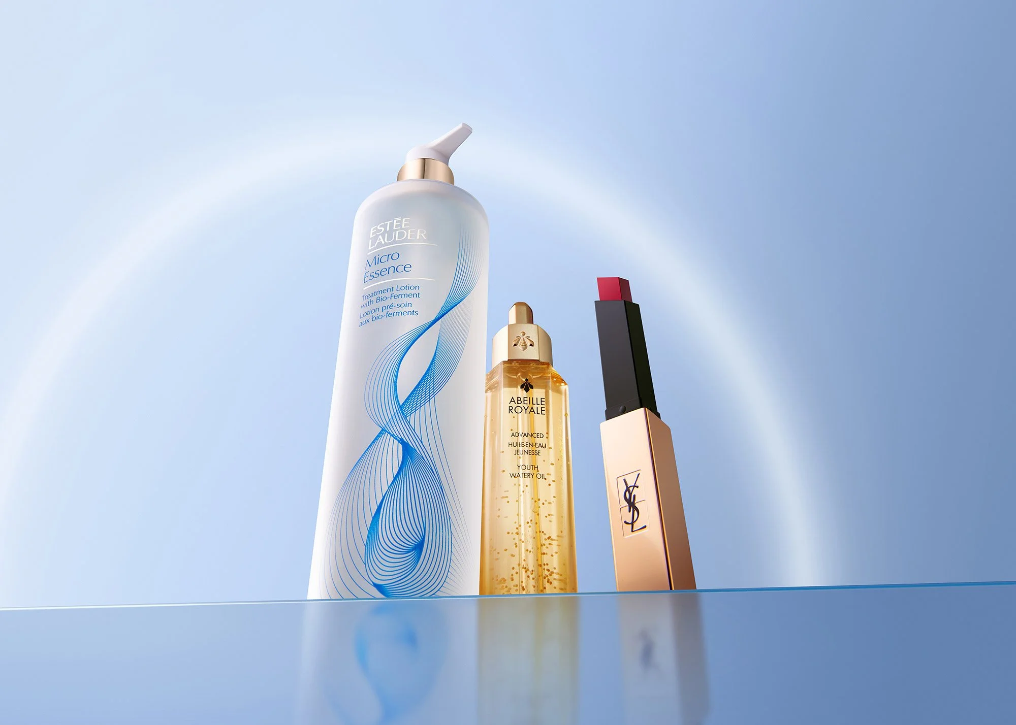 Three beauty products: a tall white bottle of Estée Lauder Micro Essence, a smaller gold bottle of Abeille Royale Youth Watery Oil, and a YSL lipstick with a red lipstick bullet. They are placed on a reflective surface against a light blue background