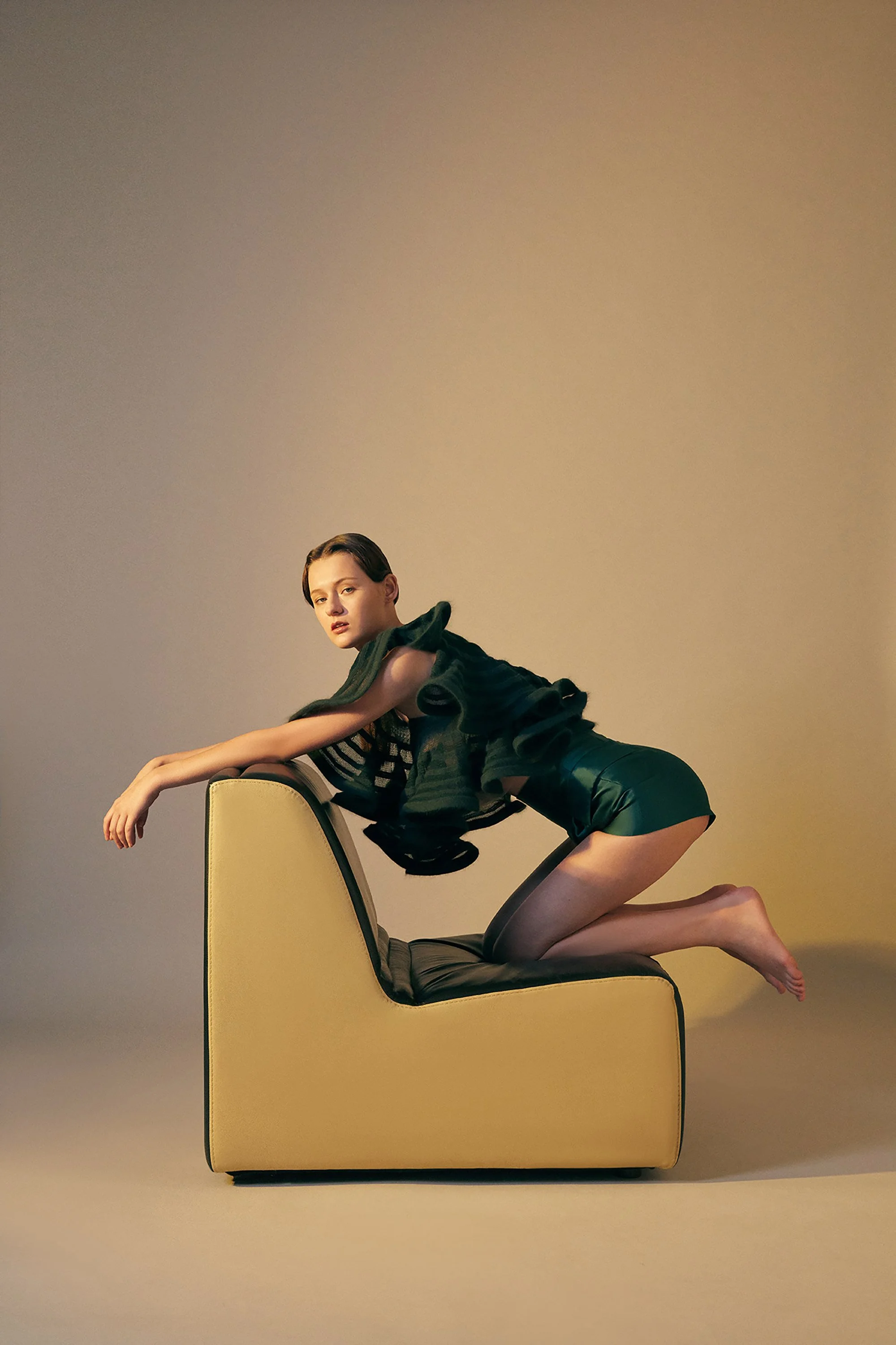 A woman with short hair in a black and green outfit kneels on a yellow and black armchair with her arms resting on the back of the chair, against a plain beige background.