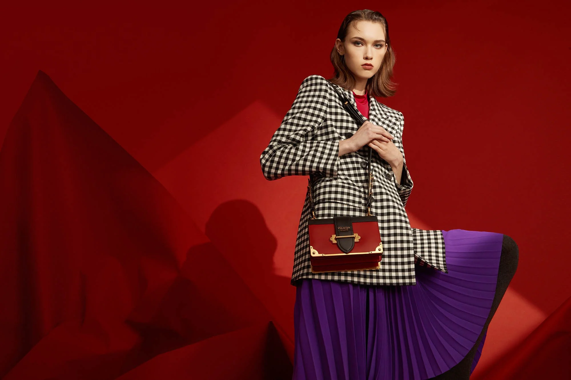 Fashion model with a black and white checkered blazer, purple pleated skirt, carrying a red Prada handbag, posing against a red background.