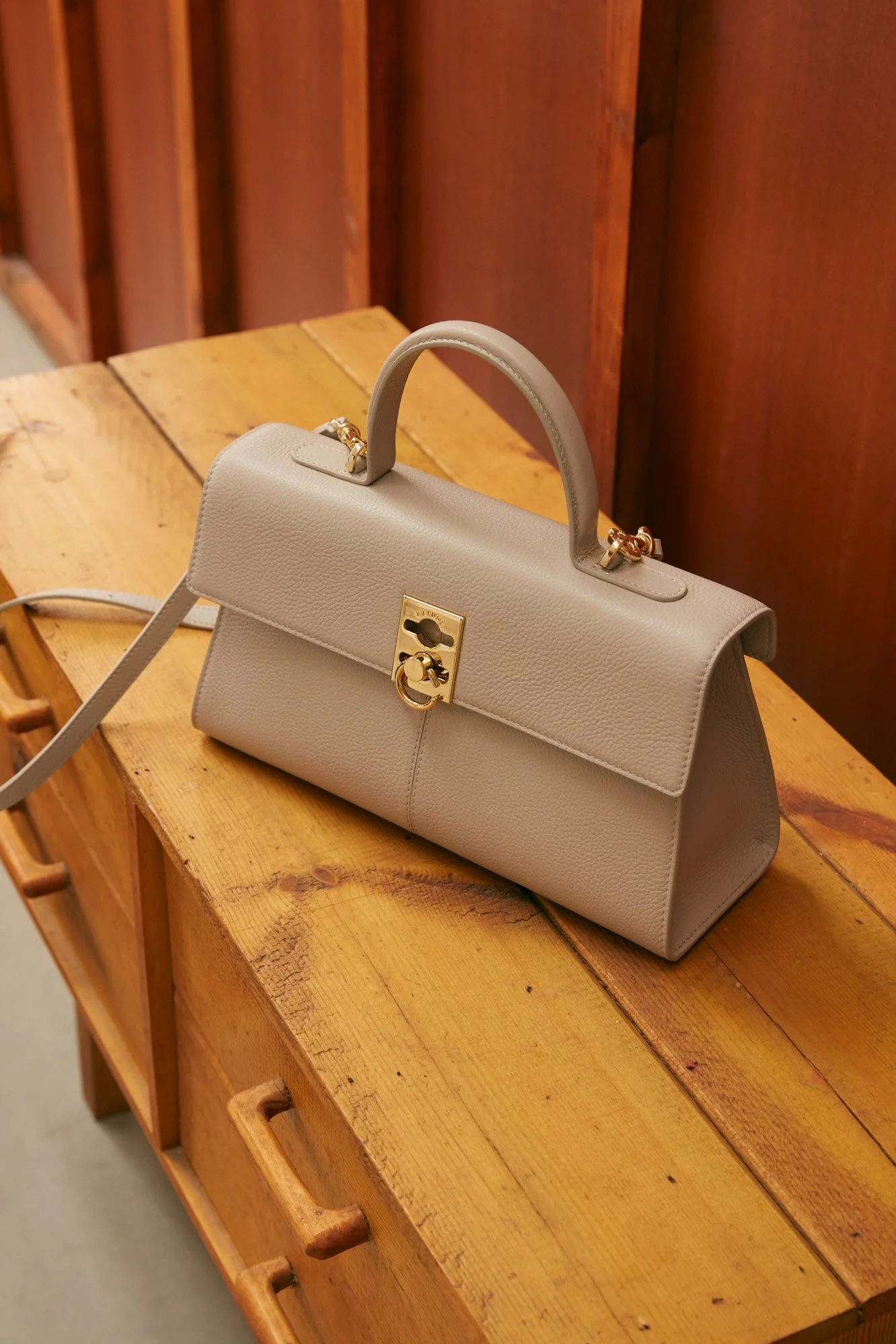 A beige leather handbag with gold hardware sitting on a wooden table with a rustic finish.