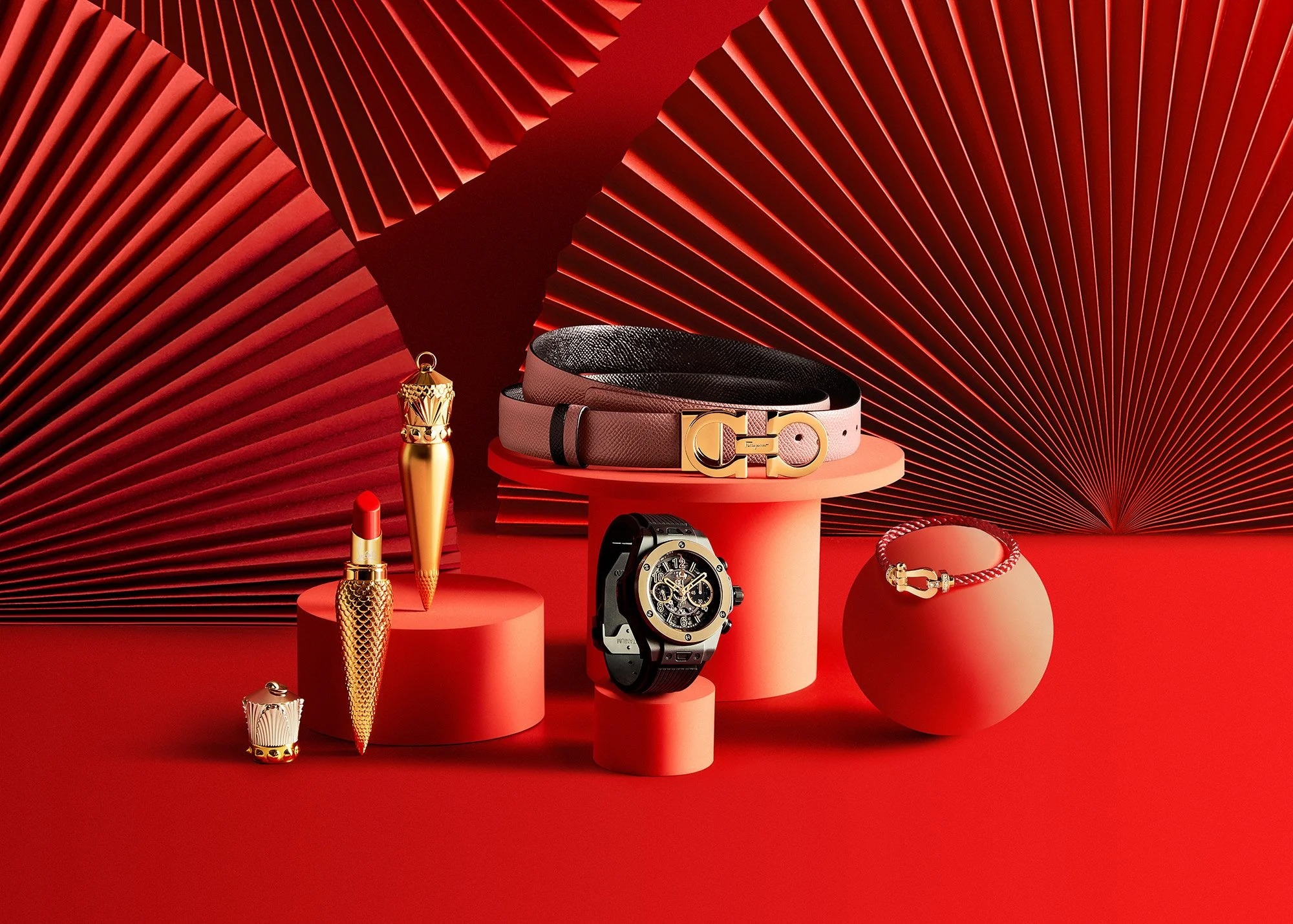 Luxurious jewelry and accessories display on red geometric background, including a black and pink belt with gold buckle, a red and gold bracelet, a black and gold wristwatch, a gold lipstick, and a gold cone-shaped ornament, arranged on pink cylindri