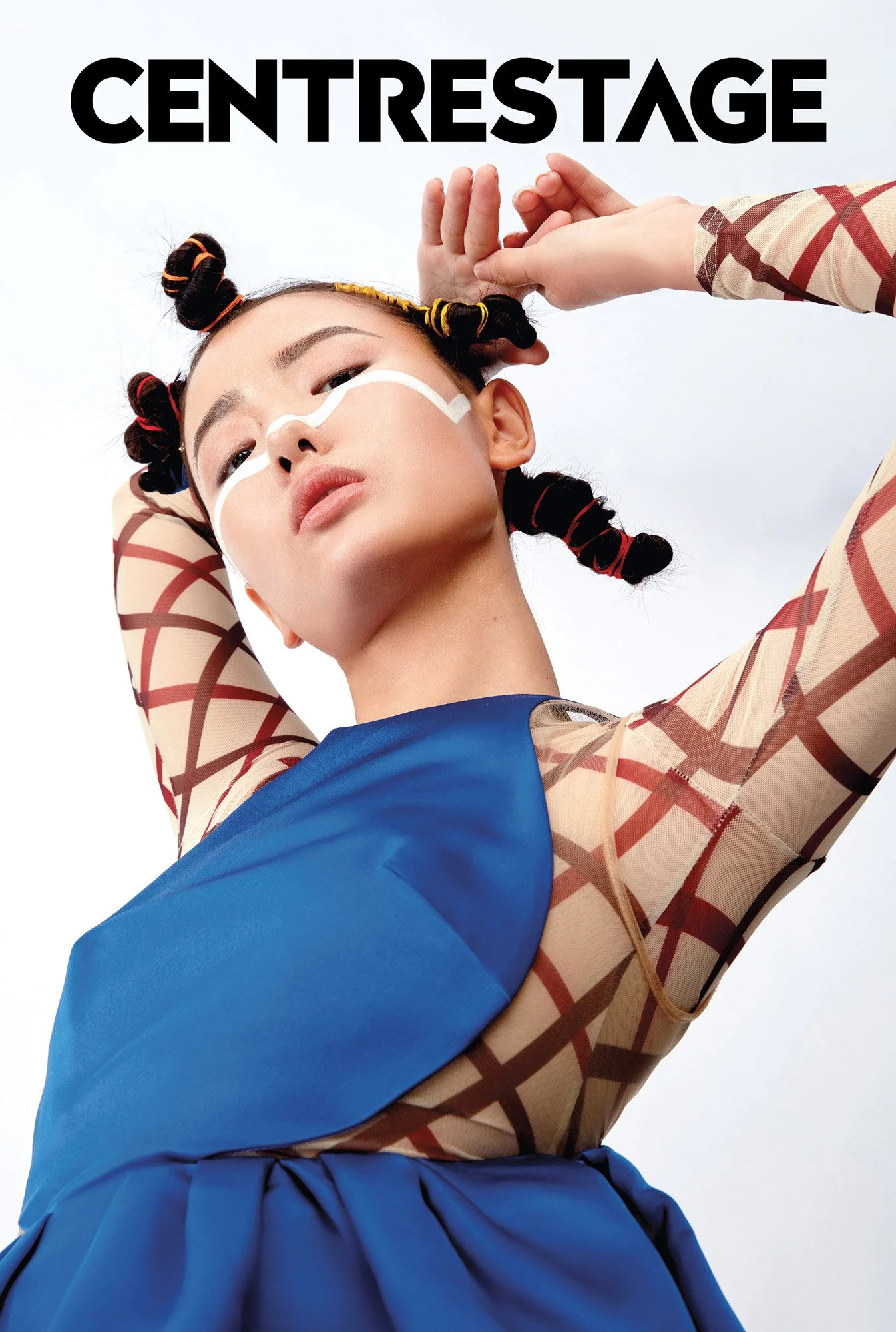 A model with styled hair in colorful curls, wearing a beige and brown patterned top with sheer sleeves, and a blue garment over her shoulders, stands against a light sky background. Her face has white makeup lines across her cheeks and nose, and she 