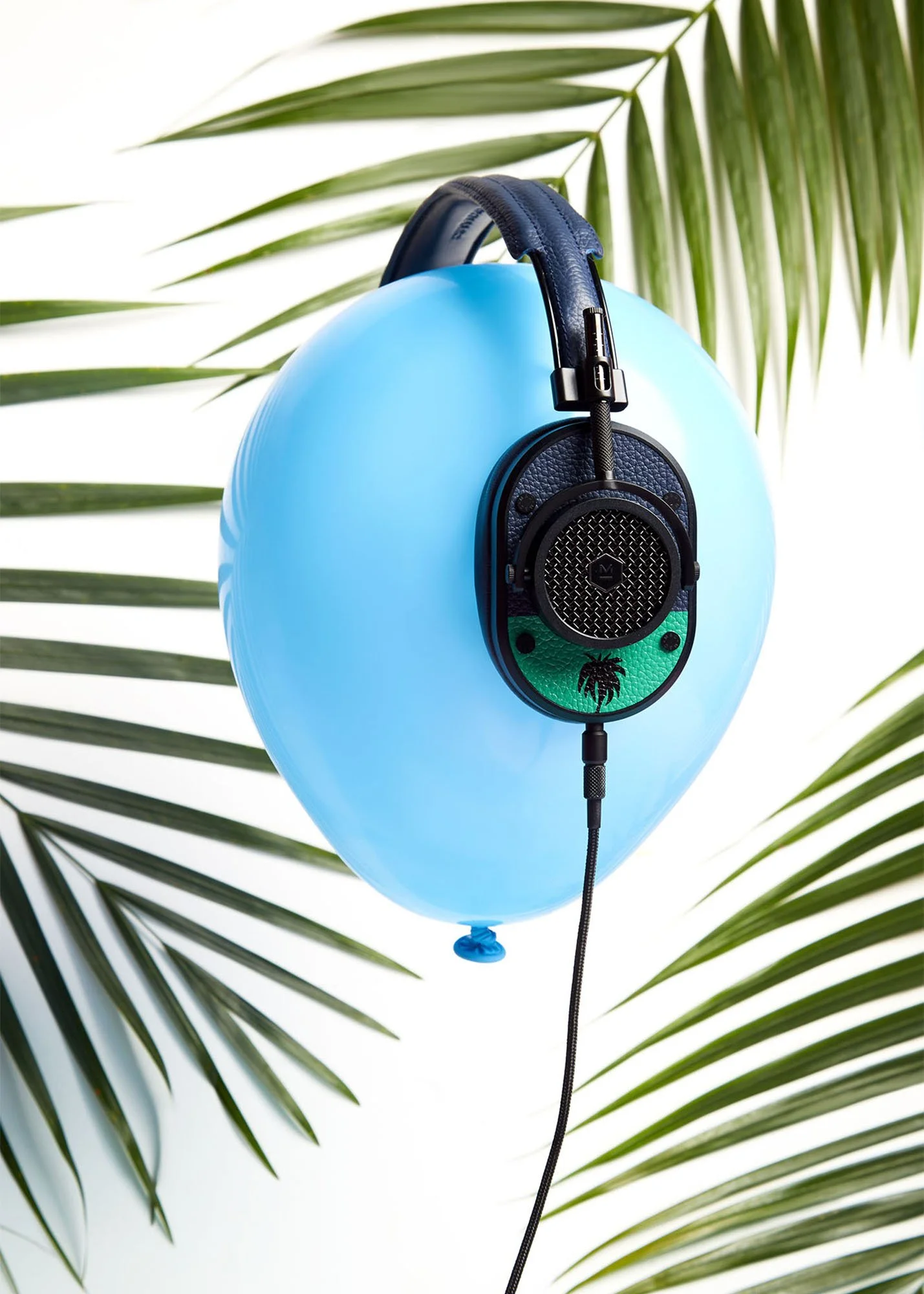 A pair of black headphones hanging from a blue balloon against a white background with green palm leaves.