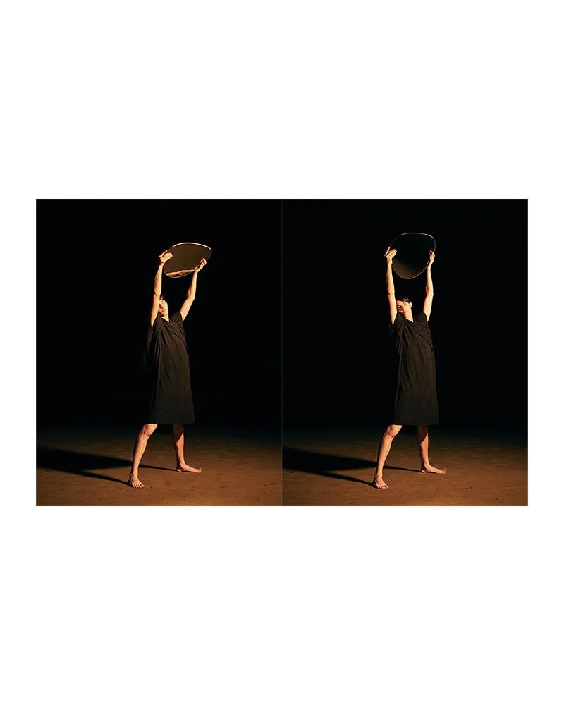 Person in black dress raising a wooden circle above their head, standing on stage with a dark background, mirrored image.