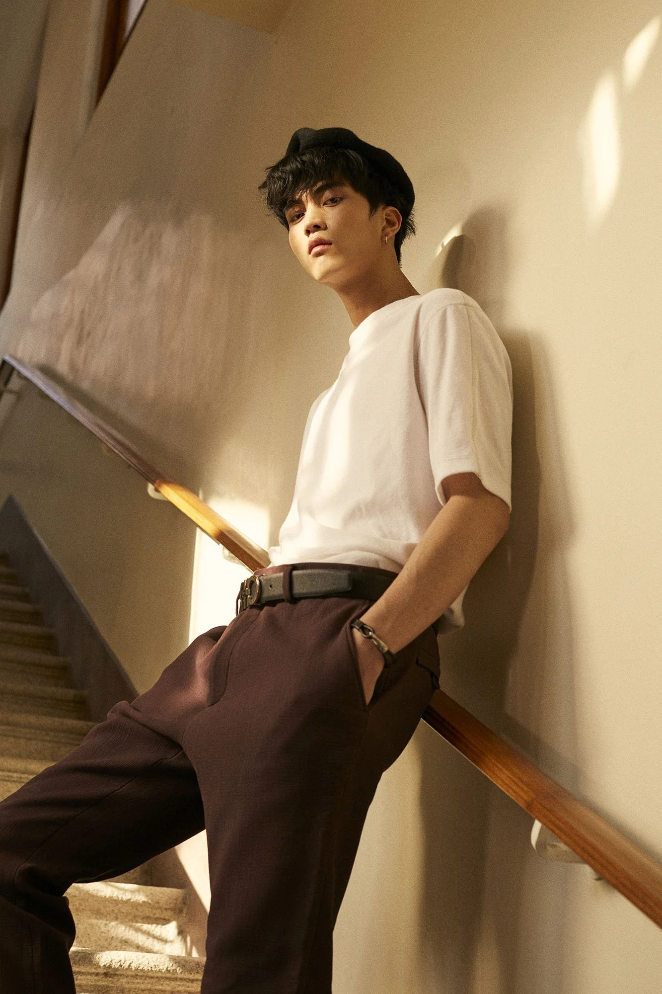 Young man with dark hair, wearing a white t-shirt, black hat, burgundy pants, and a belt, leaning against a stairwell wall, with his hands in his pockets and a watch on his left wrist.