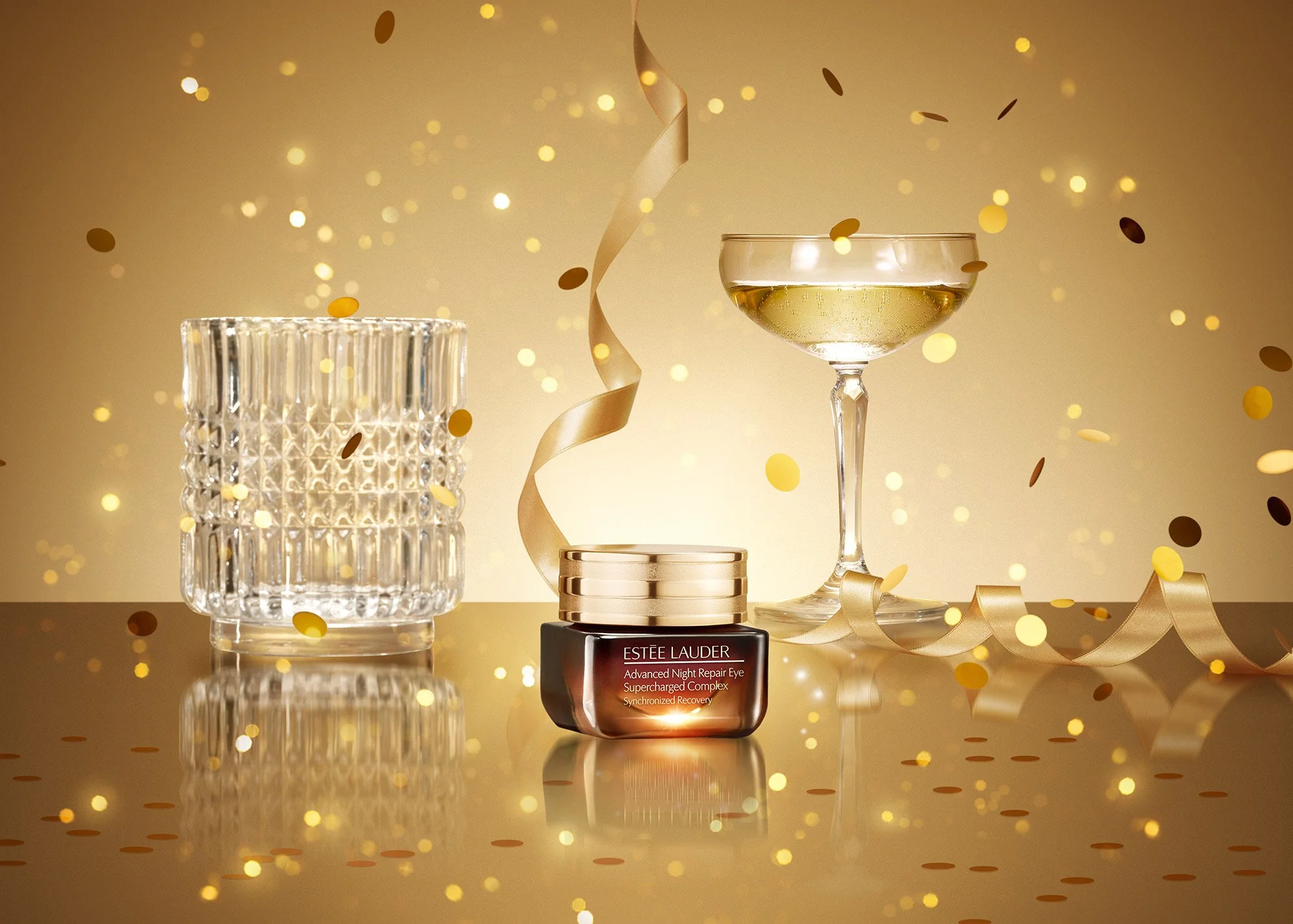 A small jar of Estee Lauder eye cream, a glass of champagne, and a decorative glass jar, with gold confetti and streamers in the background.