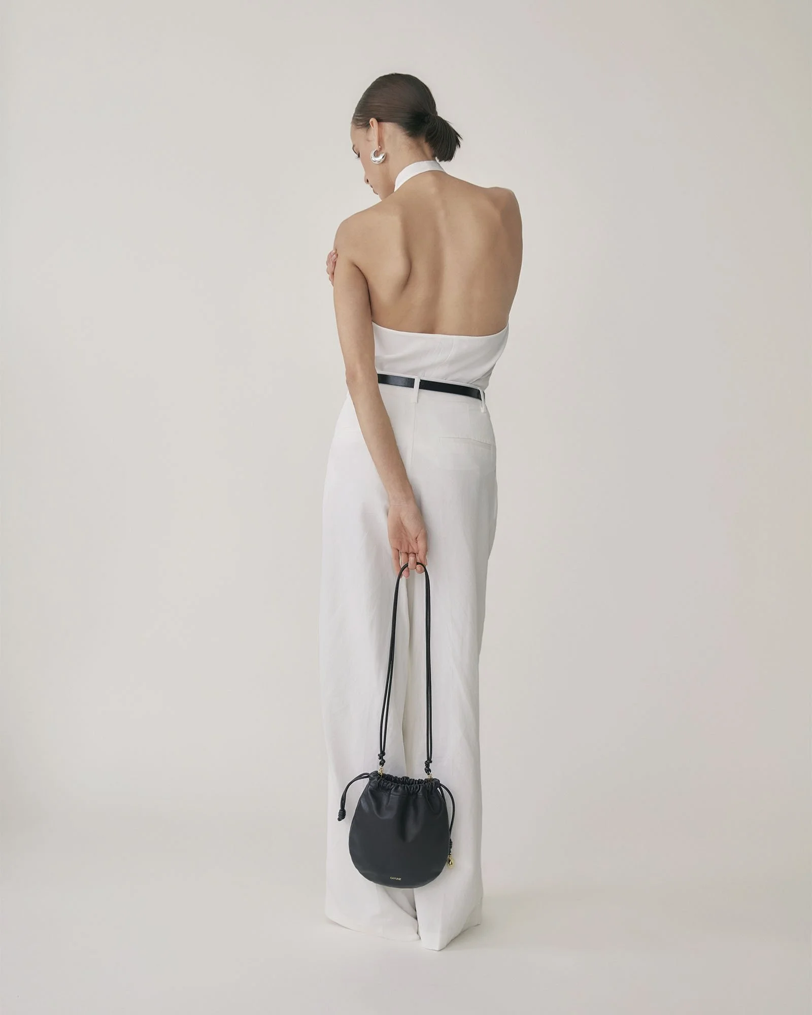 A woman with short dark hair, wearing white pants and earrings, is facing away from the camera. She is holding a small black drawstring bag with a long strap.