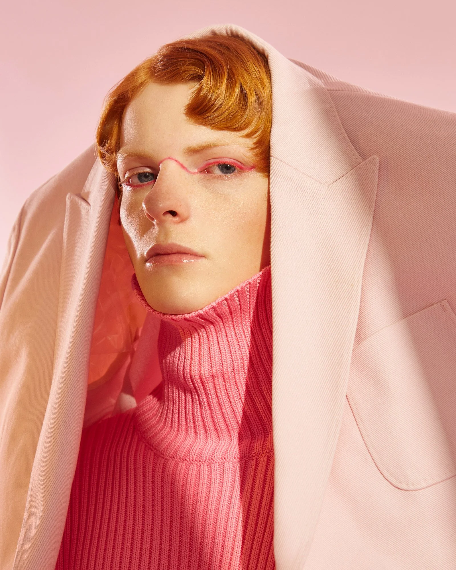 Person with light skin and red hair wearing a pink coat over a pink turtleneck against a pink background.