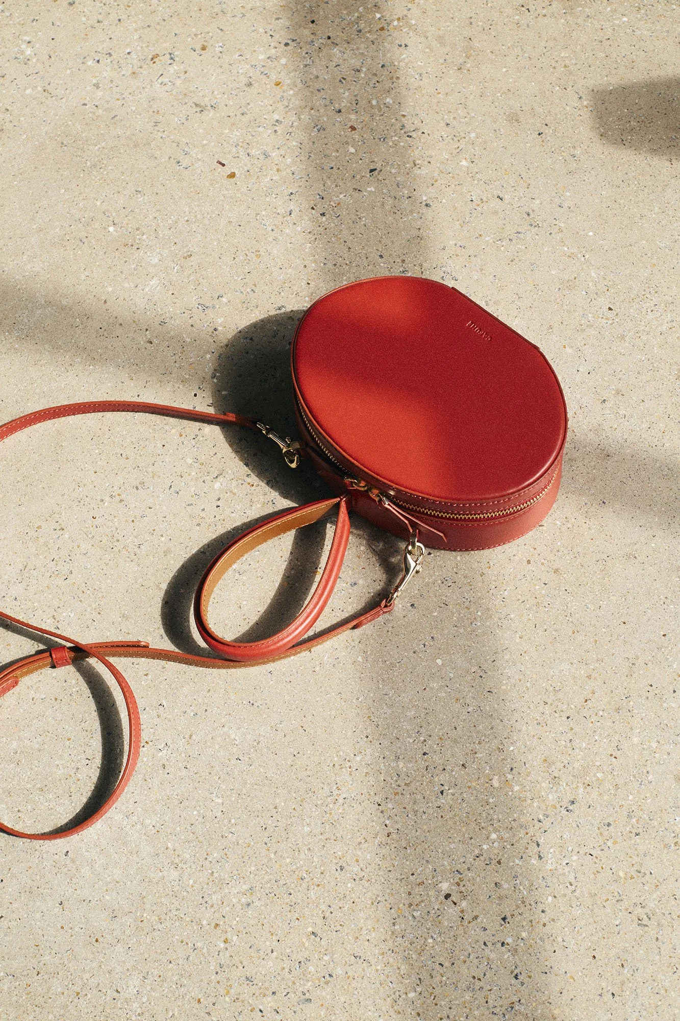 A red oval-shaped purse with a gold zipper and a long strap is lying on a sandy surface, with shadows cast by nearby objects.