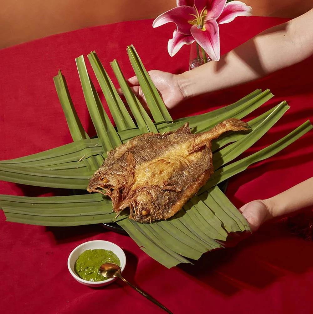 A person holding a large tropical fish, fried, on a bed of green leaves, with a flower in the background and a small bowl of green sauce.