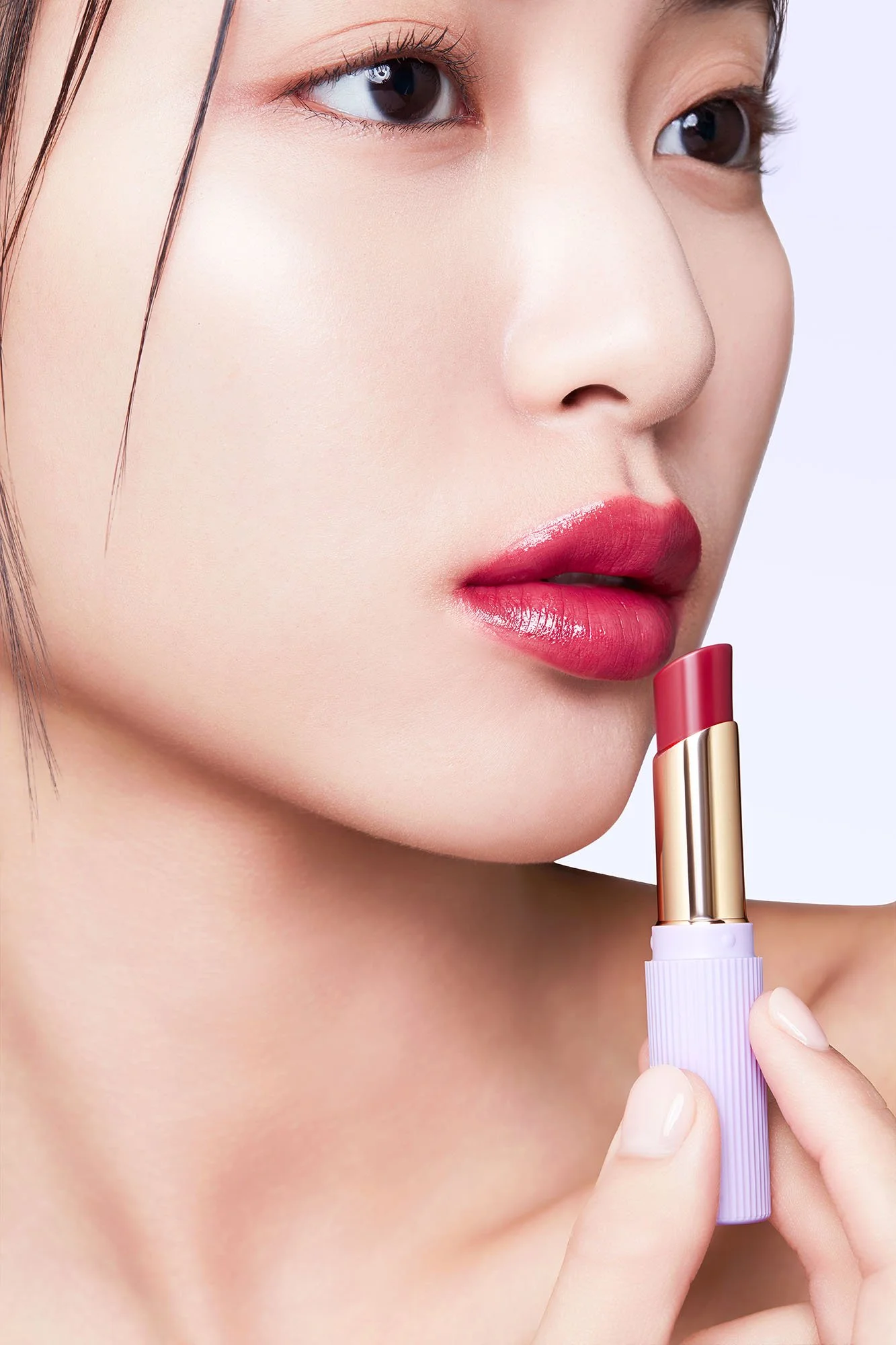 Close-up of a woman with flawless skin applying pink lipstick from a tube with lavender and gold packaging, with her face turned slightly to the side.