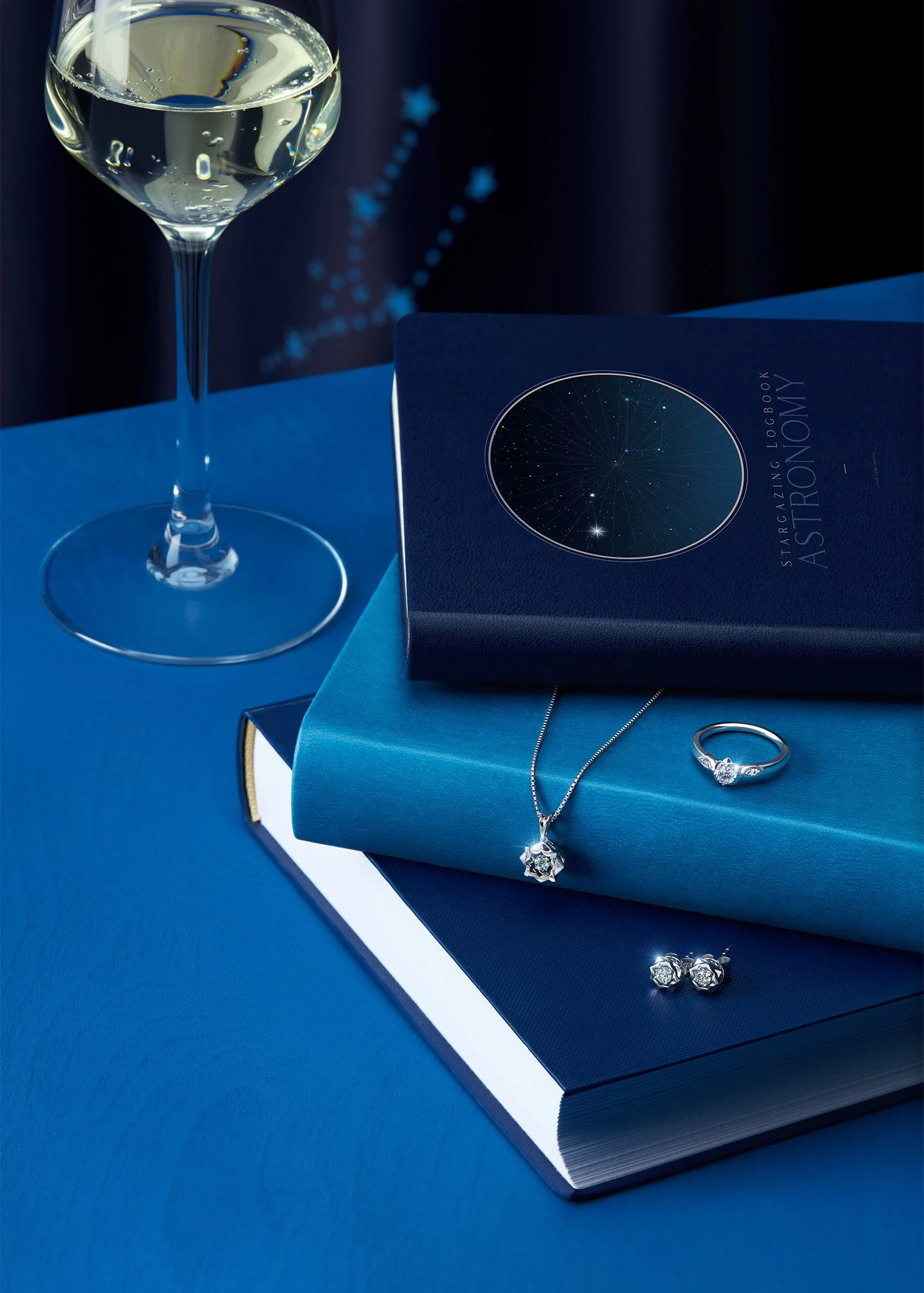 A glass of white wine, a book titled 'Star Gazing Logbook Astrology,' and jewelry including a necklace, ring, and earrings are arranged on a blue surface.