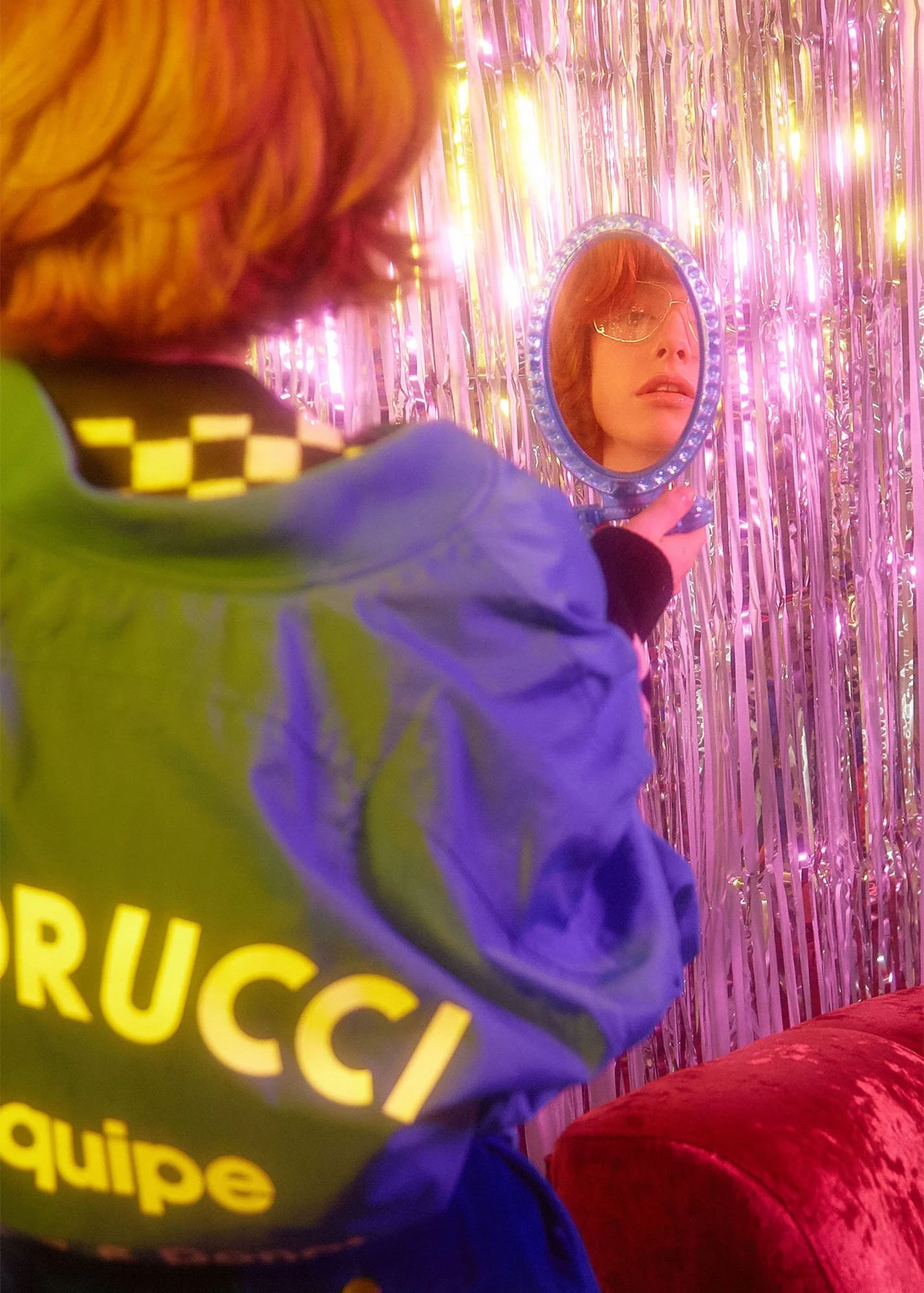 Person holding a small mirror in front of a pink metallic curtain background, wearing glasses and looking at their reflection.