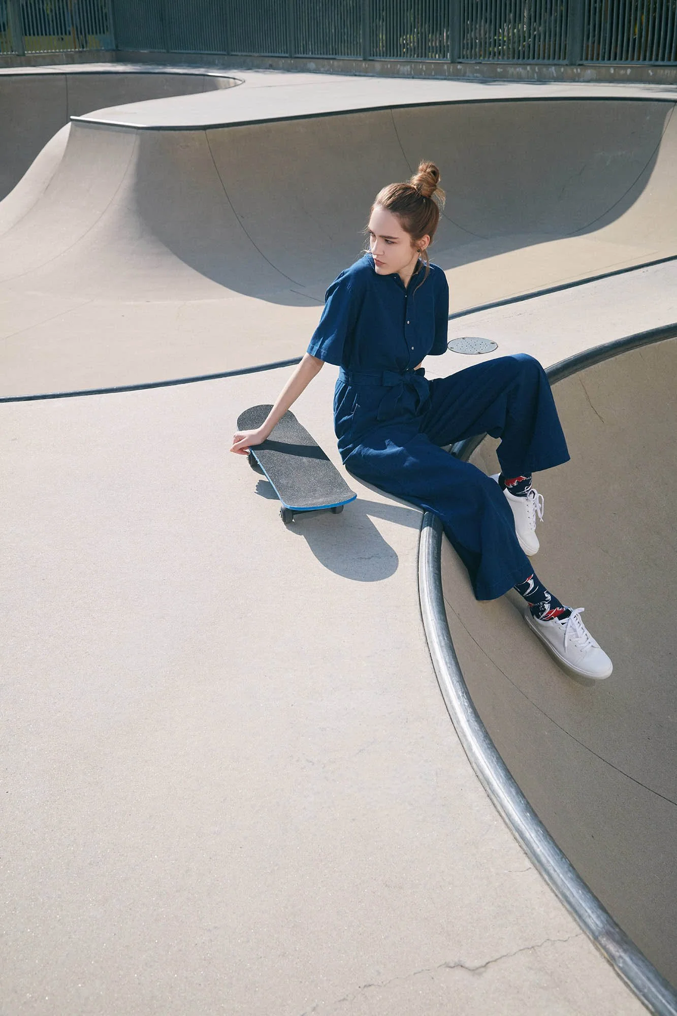 A young woman in a blue jumpsuit and white sneakers sitting on the edge of a concrete skatepark bowl with a skateboard beside her.