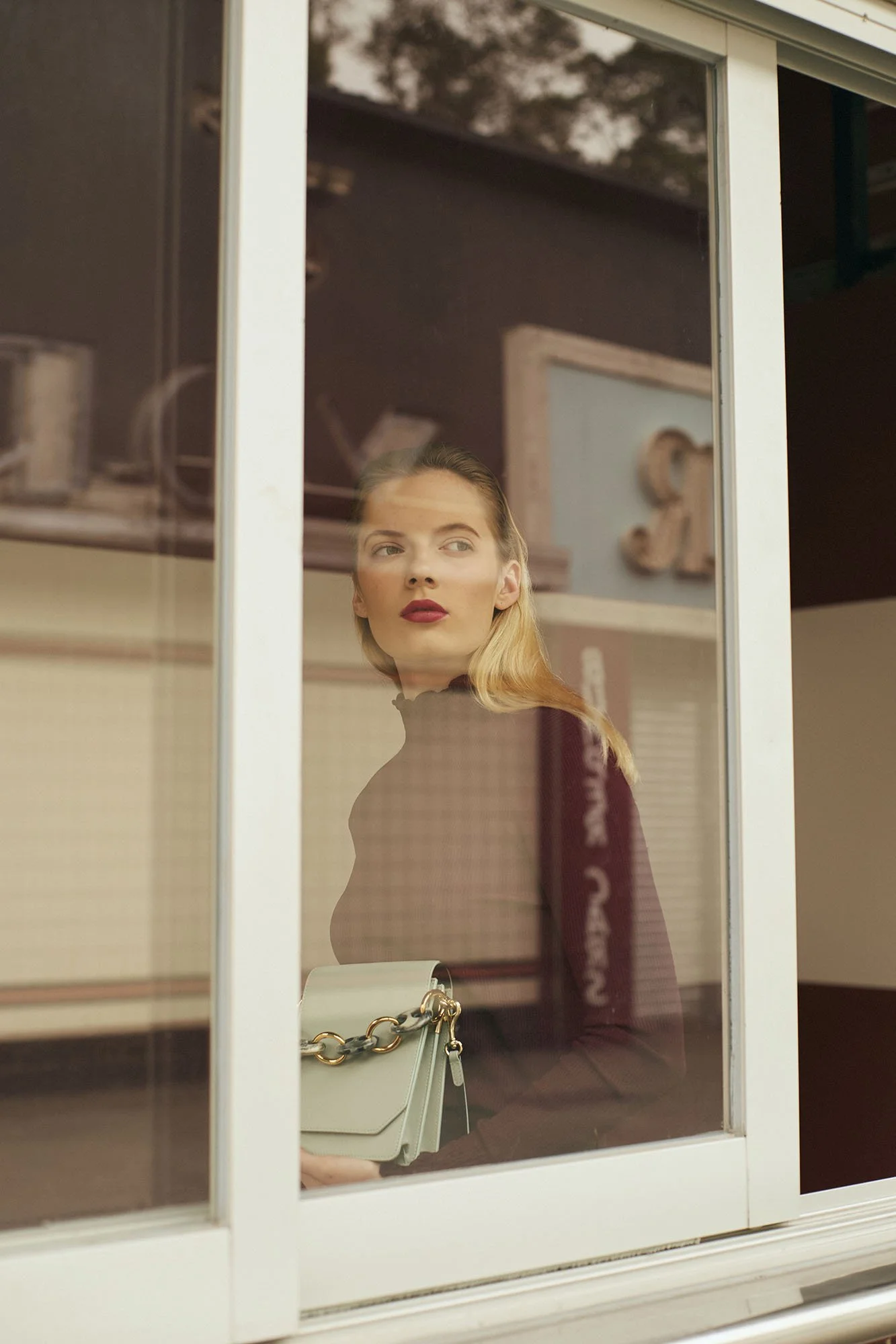 A woman with blonde hair and red lipstick looking out a window with reflections of trees, holding a beige purse with a gold chain strap inside the room.